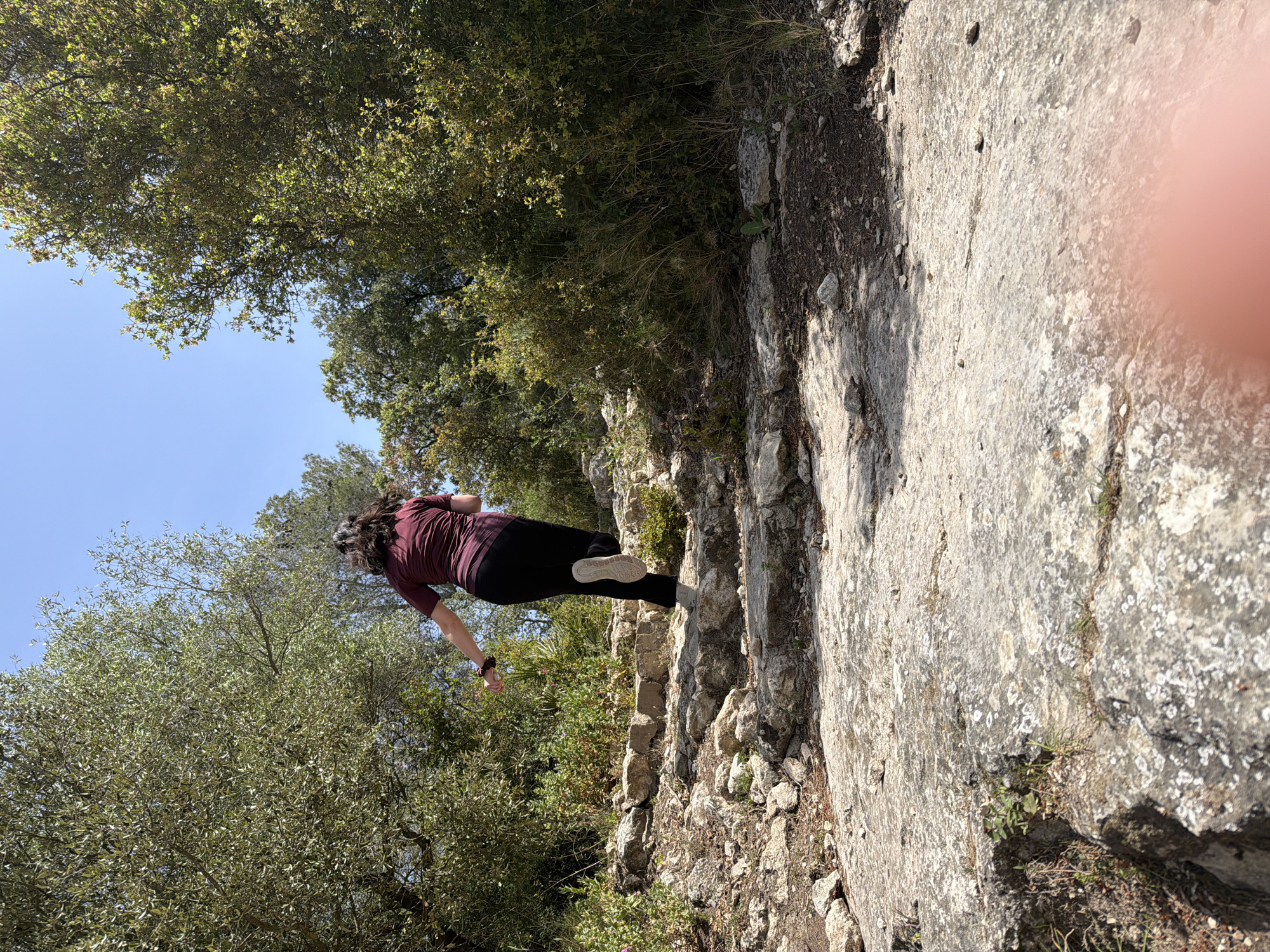 A person is captured from behind, walking or stepping carefully on a rocky, uneven path in Olèrdola, Spain. The individual wears a maroon short-sleeved top, black trousers, and light-colored sneakers. Their dark hair is pulled back. The path is made of large, light-colored rocks, some covered with lichen, and dry earth with sparse vegetation like grass and small bushes.
The scene is an outdoor, natural environment, likely a mountainous or hilly area. Dense green trees and foliage flank both sides of the path, with a clear, bright blue sky visible above and between the tree canopies. The strong sunlight suggests it is midday or early afternoon, casting distinct shadows on the rocky ground. The camera is held at an angle, making the ground appear significantly tilted. A blurry, pinkish area on the far right edge indicates a finger partially obstructing the lens. No text is visible in the image.