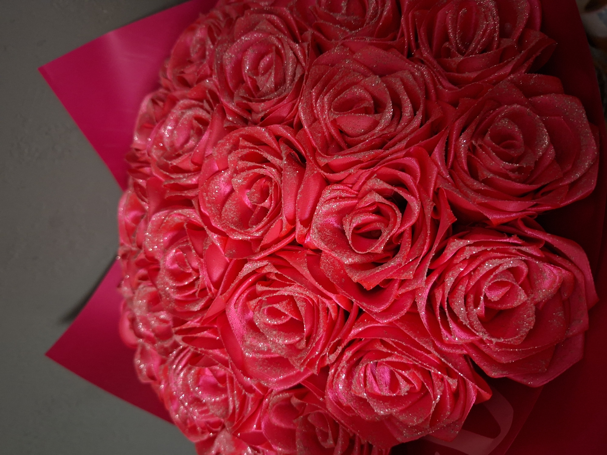A close-up shot shows a bouquet of red roses wrapped in pink paper. The roses have a slight shimmer on their petals, suggesting a light dusting of glitter.