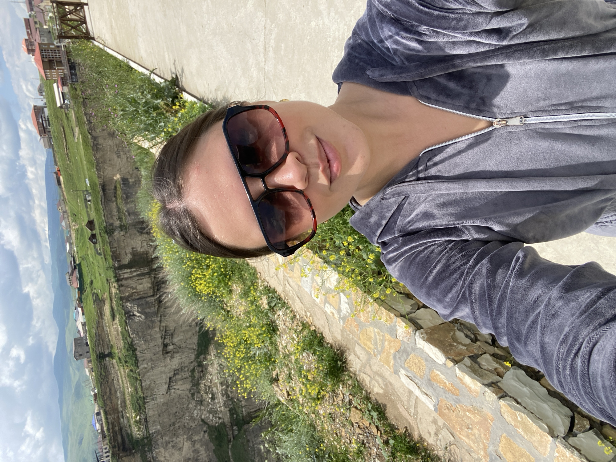 A woman in sunglasses and a grey velour tracksuit takes a selfie in front of a dramatic cliffside landscape in Khunzakh, Russia. The background features a deep canyon with sheer, rocky walls, and a village of houses perched on the opposite rim. Green hills and mountains stretch into the distance under a partly cloudy sky. Yellow wildflowers grow along the path where the woman stands. The path is paved and edged with a stone wall.