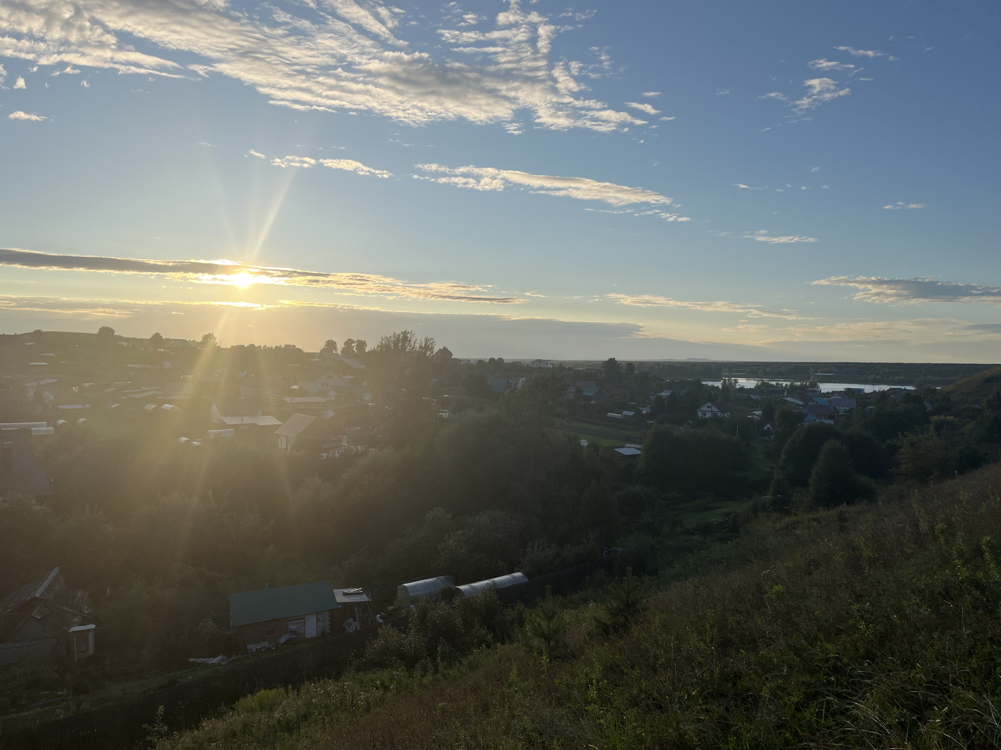 The image depicts a sun-drenched rural landscape, likely in the village of Olkhovka, Russia, during either sunrise or sunset. The low-angle sun casts long, brilliant rays across the sky and illuminates a cluster of buildings and vegetation in the foreground and middle ground. The sky is a vibrant blue, dotted with scattered white clouds, some of which are tinged with yellow and orange from the sun's light.
In the middle ground, a collection of houses with various roof colors and shapes are nestled amongst dense green trees. Some structures appear to be traditional Russian rural dwellings, while others might be more modern constructions. Several greenhouses are visible, suggesting agricultural activity. A body of water, possibly a river or lake, is visible in the far distance, reflecting the light of the sky.
The foreground consists of a grassy hillside, with tall dry grass and some scattered greenery. The overall impression is one of a peaceful, sunlit rural environment. There are no people visible in the image. The scene is static and captures a moment in time.