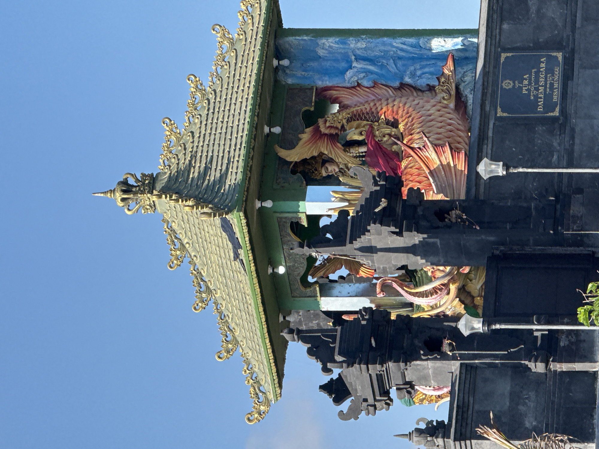 The image shows a portion of the Pura Dalem Segara temple in Desa Munggu, Mengwi, Indonesia. The temple structure is elaborately decorated with carvings of mythical creatures, including a large, ornate fish-like figure with red and orange scales. The roof is multi-tiered with a green hue, adorned with golden ornamental edges. A black sign displaying the temple's name is visible. The sky above is a clear blue. The image is taken from a low angle, looking upwards.
