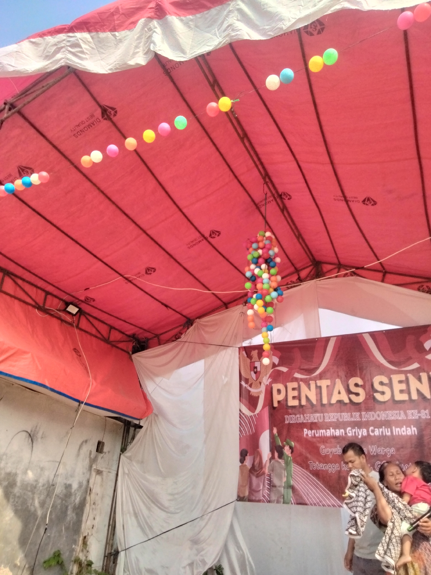 The image depicts an outdoor event under a red tent. The tent is supported by metal poles and has white trim along its edge. Strings of colorful, round decorative lights are strung across the tent, with a large cluster of these lights hanging centrally. In the background, a banner reads "PENTAS SEN...", with smaller text below indicating "DIRGAHAYU REPUBLIK INDONESIA KE-81", "Perumahan Griya Carlu Indah", and "Tetangga Ke...". This suggests the event is taking place in Carlu Housing, Griya, in Cariu, Indonesia, celebrating Indonesia's 81st Independence Day.
A man and a woman are in the foreground, with the woman holding a young child. The woman is looking up and appears to be singing or speaking, with her mouth open. The man is looking down towards the child. The backdrop behind them is a white drape, partially obscuring the banner. The environment appears to be somewhat rustic, with a textured wall and some greenery visible on the left side. The overall scene suggests a community celebration or performance.