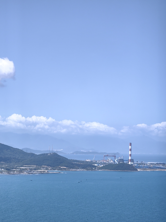 A panoramic view features a blue ocean under a vast sky with scattered clouds. Distant mountains fade into the horizon, partially obscured by a layer of low-lying clouds. A cluster of industrial buildings and a tall chimney with red and white stripes are visible on a peninsula extending into the sea. Several small boats are scattered on the water.