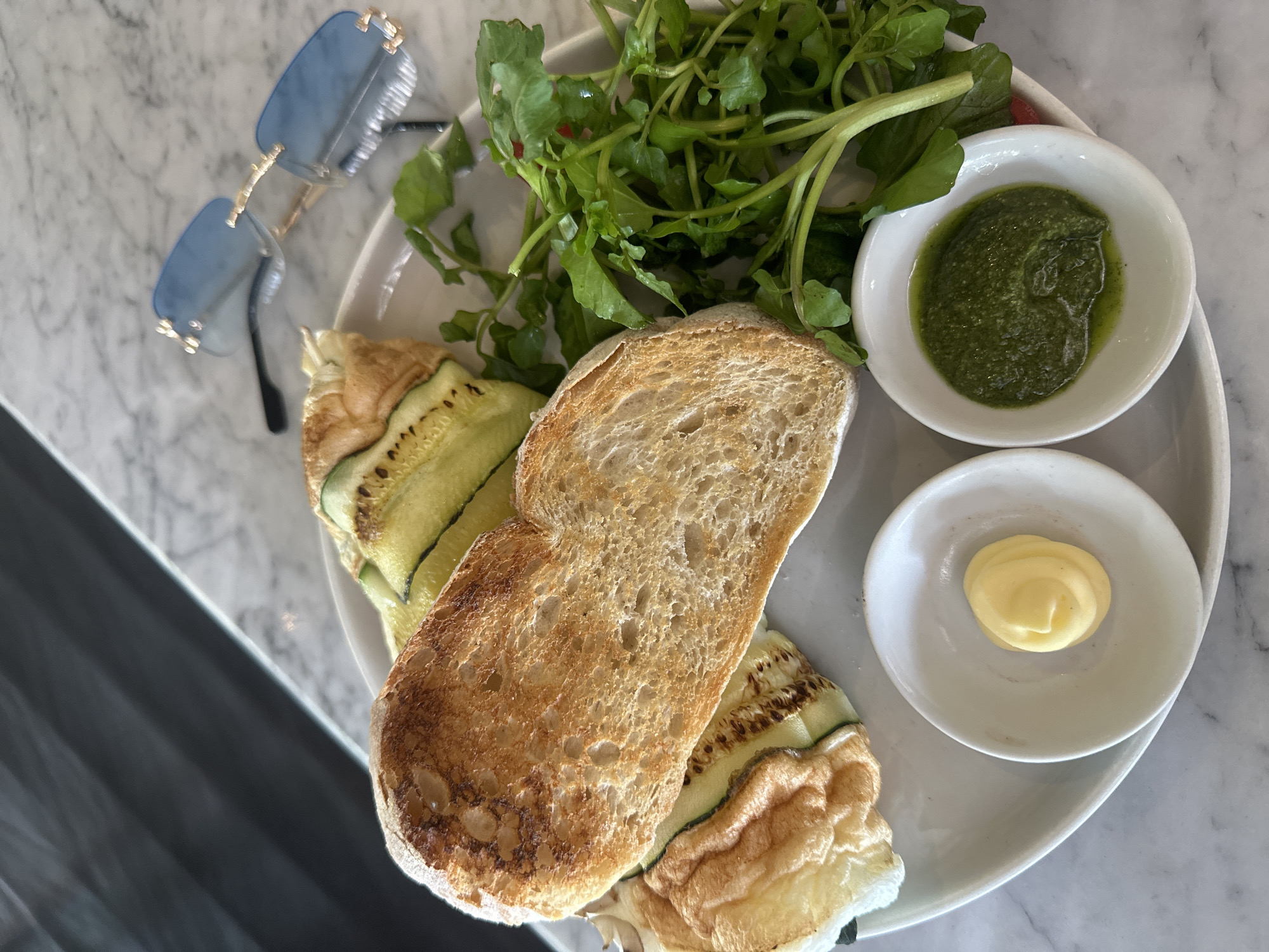 On a white marble tabletop in North Kuta, Indonesia, a white plate holds a meal. A toasted bread slice rests on grilled zucchini. Green watercress is arranged to the side, along with two small white bowls: one filled with green pesto, the other with yellow butter. A pair of light blue sunglasses with gold frames sits to the upper left of the plate. The scene is brightly lit, possibly outdoors.