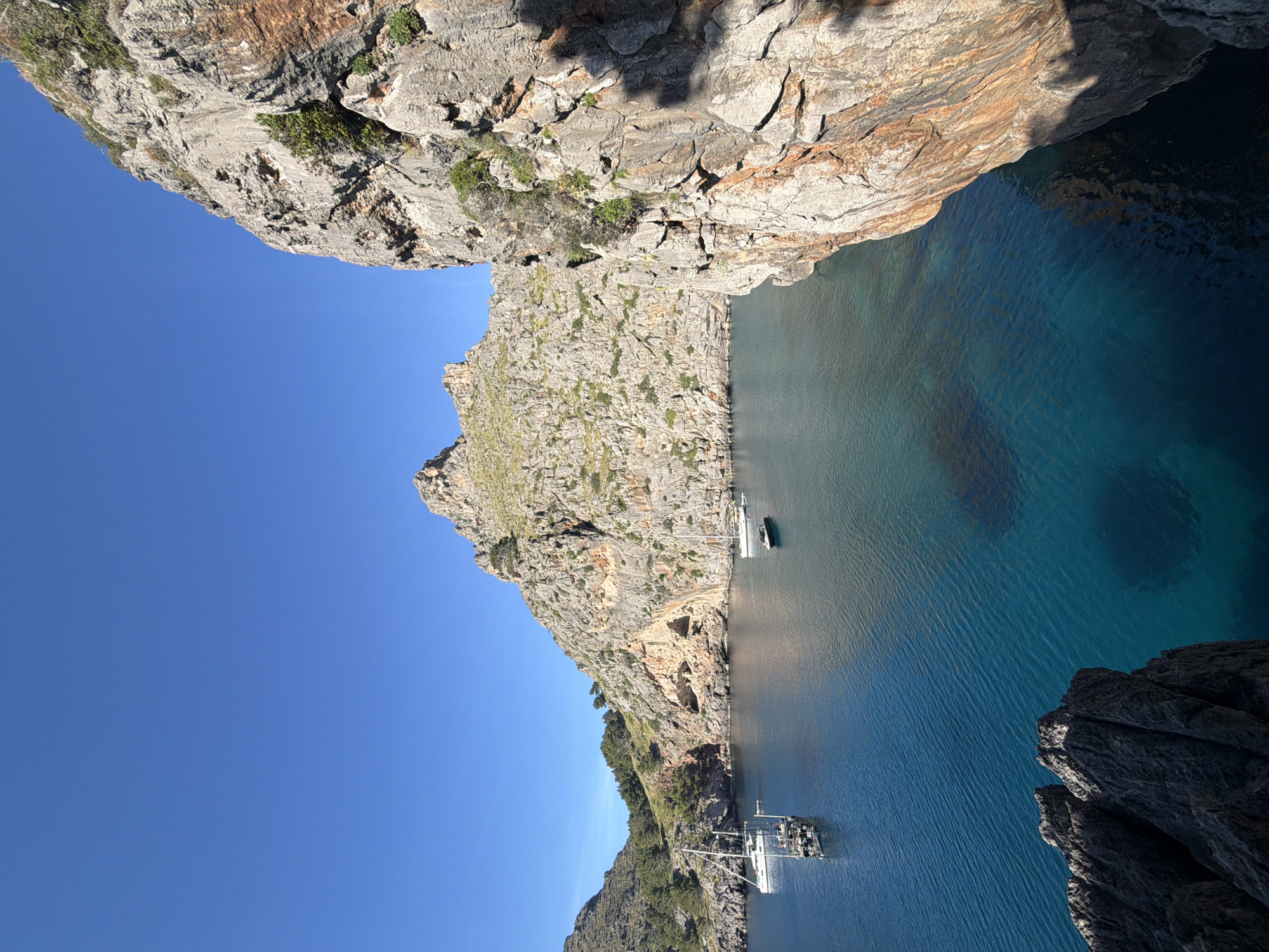 The image shows a view of the Torrent de Pareis gorge in Escorca, Spain. The gorge is a narrow inlet of the sea, surrounded by tall, rocky cliffs on both sides. The water is deep blue and clear, reflecting the sky. There is a yacht and a smaller boat in the gorge. The sky is cloudless and blue. The cliffs are mostly gray and brown with some vegetation growing on them.