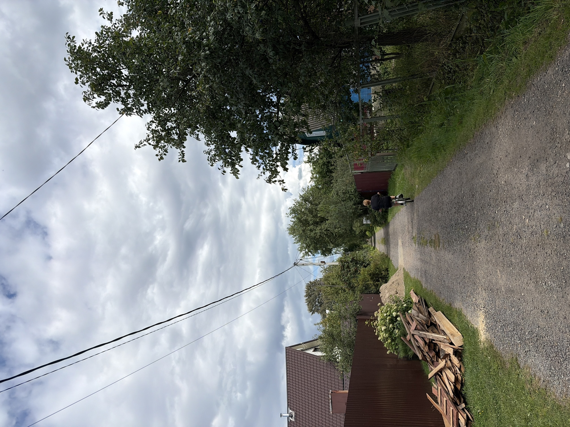 The image depicts a gravel road in a rural or semi-rural area, likely within the "Керамик Снт (белая, Russia)" location. The road is flanked by vegetation and fences, with houses and trees visible on either side. In the foreground, a pile of lumber and sand sits on the left side of the road. Further down the road, a person is riding a bicycle away from the camera. Overhead, power lines stretch across the sky, which is filled with cloudy formations. The scene suggests a quiet, possibly residential, setting on a day with overcast weather.