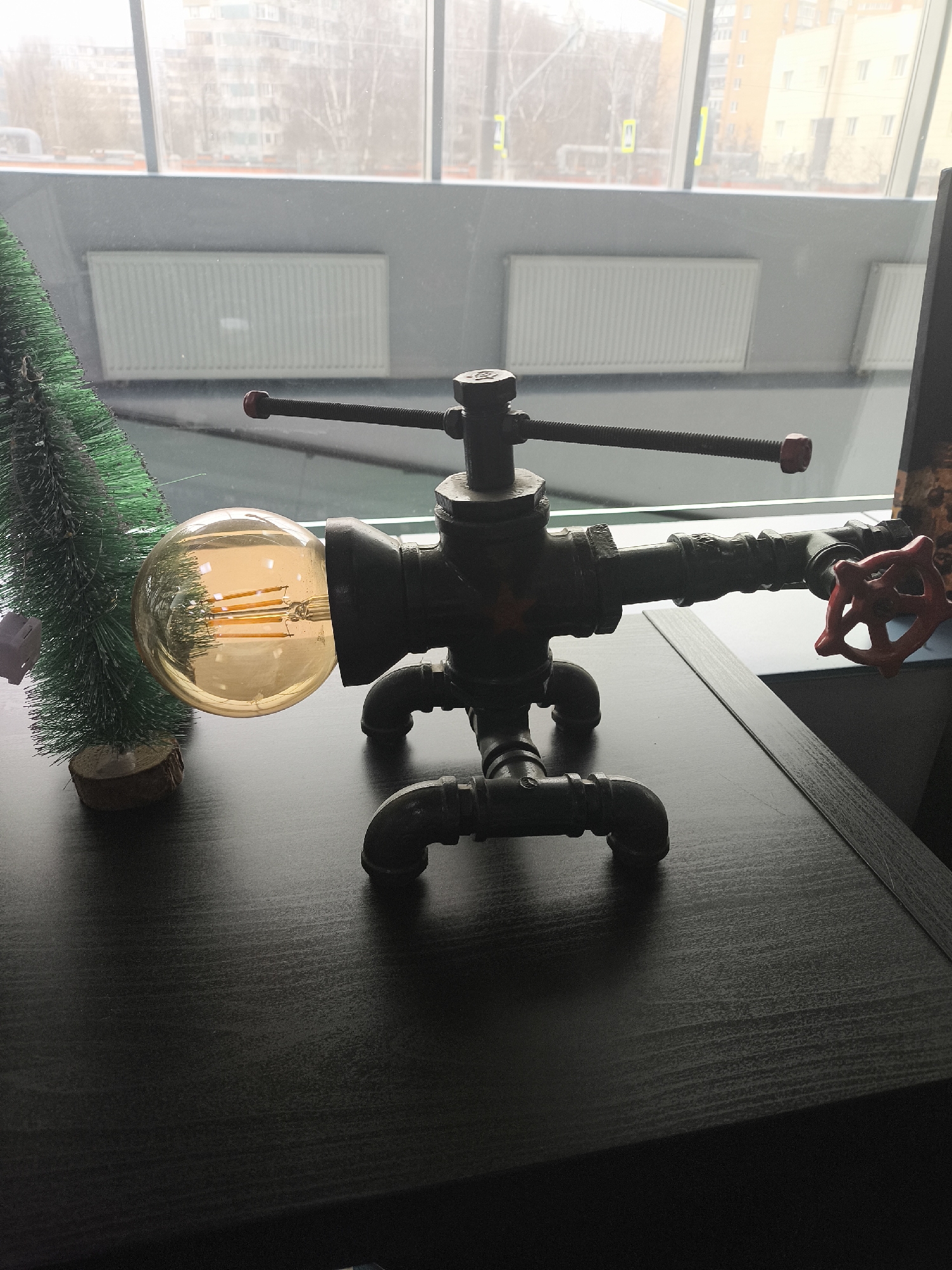 On a dark, wood-grain surface rests a steampunk-style lamp made of pipes and fittings. The lamp features an Edison-style bulb and a red valve as a switch. To the left of the lamp is a miniature Christmas tree. The scene is set indoors, with a large window in the background revealing a cityscape with buildings and bare trees. The location is an urban area, city of Unknown, Unknown.