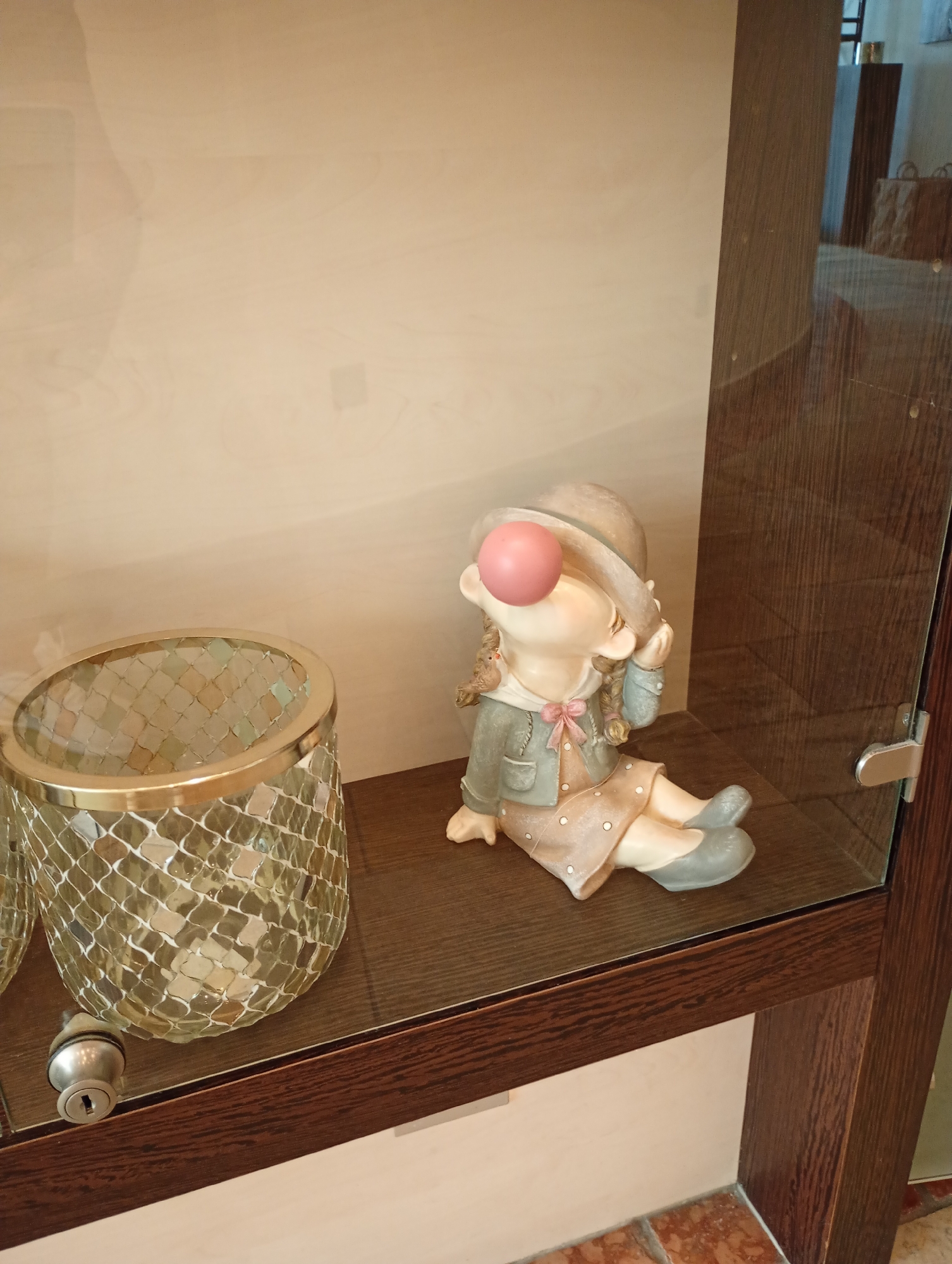 A figurine of a young girl wearing a hat, jacket, and dress sits on a wooden shelf inside a glass display case. The figurine has a pink ball on its nose, and its right hand is near its face. To the left of the figurine is a decorative glass vase with a gold rim. A silver lock is visible on the shelf in front of the vase. The background of the display case is a light-colored wall. The scene is set in the city of Bad Sachsa, Germany.