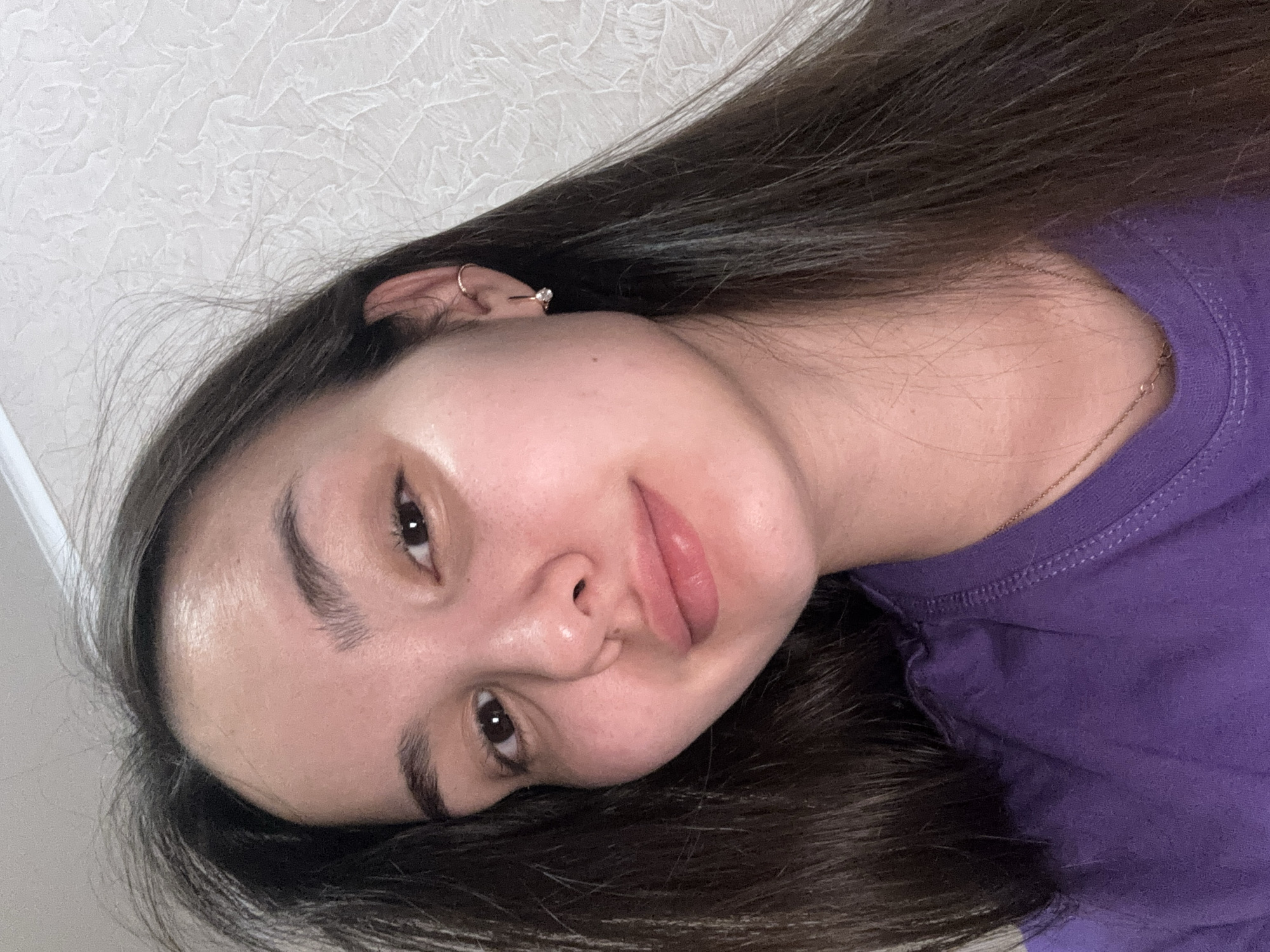 A young woman is depicted in a close-up, headshot-style photograph, lying down with her head tilted, looking directly at the camera. Her long, dark brown hair is spread around her head, framing her face and covering parts of the image. She has a relaxed and natural expression, with dark eyes and lips slightly parted in a soft pout. Her skin appears clear and even-toned, with a subtle highlight visible on her forehead and cheekbones.
She is wearing a casual purple t-shirt, visible on the right side of the frame. A thin, delicate gold chain necklace is faintly visible around her neck, peeking from beneath her t-shirt. Her left ear, partially visible, features a small gold hoop earring in the helix and a stud earring with a clear stone in the lobe.
The background is a light-colored wall with a subtle, textured pattern, suggesting an indoor setting. The lighting is soft and appears to be natural or evenly diffused, creating a serene and candid atmosphere. There is no visible text in the image. While the provided context indicates the location is Orenburg, Russia, this is not visually evident from the photograph itself.