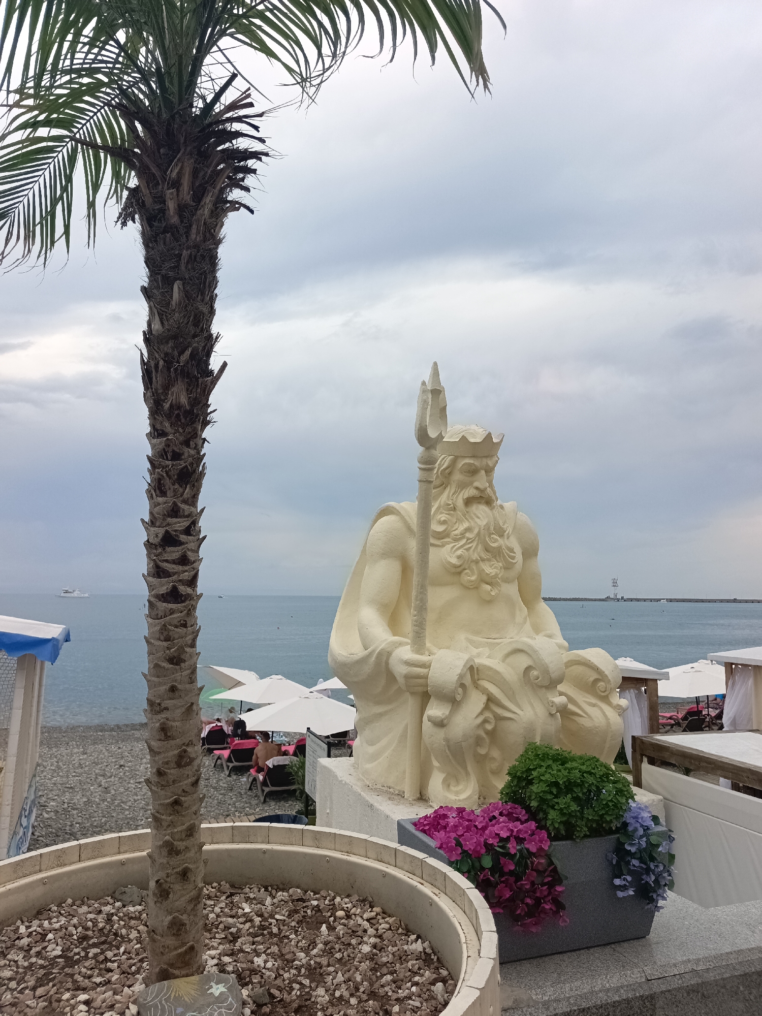 This image depicts a statue of a male figure, likely Poseidon or Neptune given the trident he holds and his regal crown, seated in front of a body of water. The statue is light-colored and detailed, with a flowing beard and muscular physique. He is positioned on a ledge, with a planter containing vibrant pink, purple, and blue flowers to his right.
In the background, the scene suggests a beach resort. A tall palm tree stands to the left of the frame, its trunk textured and weathered. Beyond the statue and palm tree, a pebble beach is visible, with lounge chairs and white umbrellas set up. Several people are seated on the lounge chairs, some appearing to be sunbathing or relaxing. The sea stretches out to the horizon, with a white boat visible in the distance and a lighthouse structure on the far right. The sky is overcast with heavy, grey clouds, indicating a potentially moody or stormy weather.
This scene is characteristic of the coastal areas of Sochi, Russia, known for its subtropical climate and seaside resorts. The presence of palm trees, a pebble beach, and the general setup of beach amenities are consistent with the resort atmosphere found in cities like Sochi. The overall impression is one of a tourist destination by the sea, with a prominent classical-style statue as a focal point.