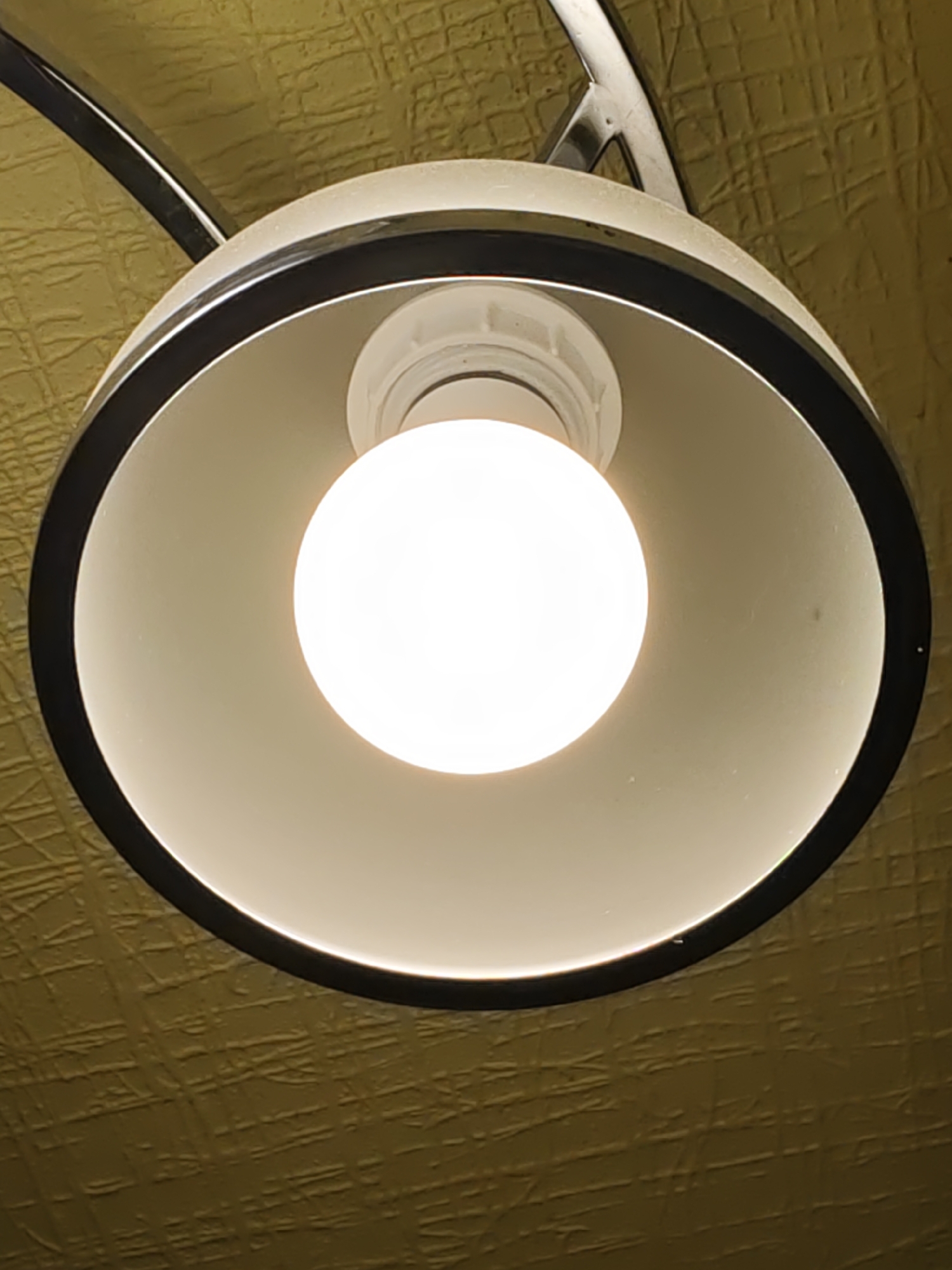 A close-up, interior, low-angle shot shows the underside of a modern ceiling light fixture against a textured yellow-gold ceiling. The fixture has a wide, shallow bowl shape with a matte white interior and a thick black trim around the rim. At the center of the bowl is a bright white, rounded LED light bulb, illuminating the inside of the fixture. A thin, metallic arm extends from the top of the fixture upwards, likely for mounting. The ceiling has a repeating, embossed pattern that resembles a grid or criss-cross texture. The image appears to be taken in Rostov-on-Don, Russia.
