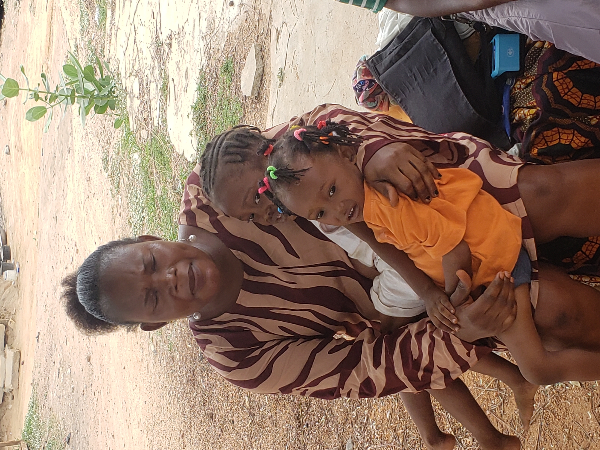 A vibrant outdoor image captures an adult woman and two young children in a warm embrace, set against a backdrop of a dirt ground with sparse vegetation, likely in Adankolo, Nigeria. The woman, with dark skin and hair pulled back in a bun, is positioned towards the left. She wears a long-sleeved dress with a distinctive brown and beige zebra-like stripe pattern and pearl earrings. Her eyes are partially closed in a relaxed expression, and she has a gentle smile.
Nestled in her arms is a toddler, also with dark skin, wearing an orange t-shirt and blue shorts. The toddler's braided hair is adorned with multiple colorful hair ties, and she looks directly at the camera with a soft smile. Partially visible behind the toddler, and held by the woman, is an older child with braided hair, wearing a striped top similar to the woman's. This child is also looking towards the camera.
The background reveals a simple, dry outdoor environment with patches of grass and a small green plant in the upper left. In the upper right corner, parts of other individuals or objects are visible, including a glimpse of a person wearing a green and white striped garment and black trousers. A black bag and a light blue rectangular object are also present, with the letters "UN" faintly visible on the blue object. The natural lighting suggests a daytime scene, conveying a sense of peace and affection within the group.
