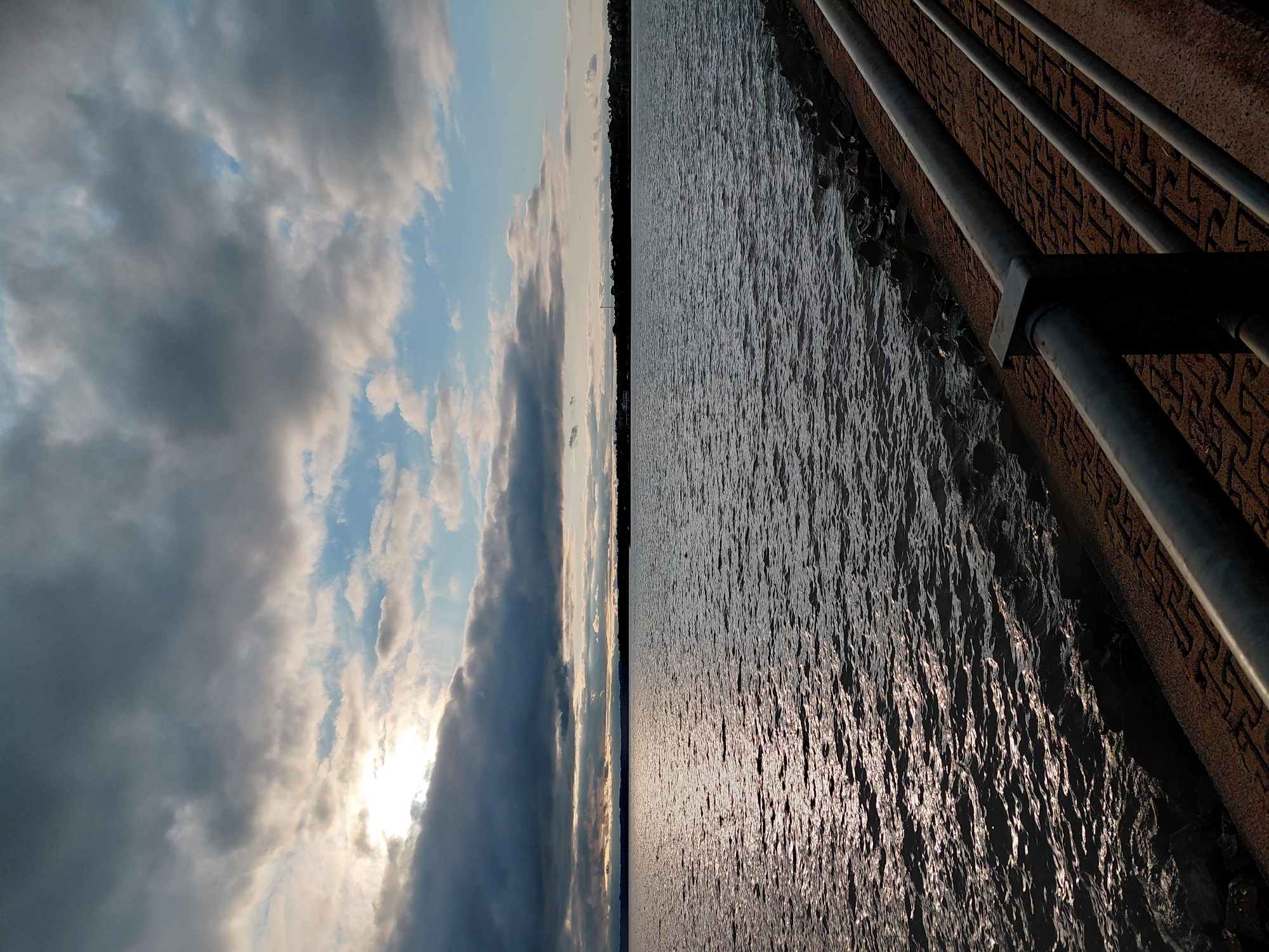 The image captures a view of the Parana River in Encarnacion, Paraguay, at dusk. The sky is filled with dramatic, heavy clouds, with patches of blue visible and the sun peeking through on the left, casting a shimmering reflection on the water. The river's surface is textured with small waves, reflecting the muted sunlight. Along the right side of the image, a paved walkway borders the river, separated by a metal railing. The walkway is made of reddish-brown interlocking pavers, and the railing is dark gray. Rocks and vegetation are visible along the river's edge. The far bank of the river is lined with dark silhouettes of trees and buildings, indicating the presence of the city. There are no people or animals visible in the frame.