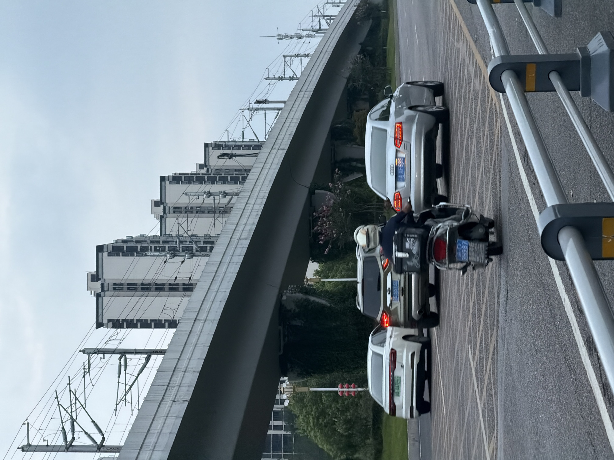 An urban scene in Jiangjin, China, features a multi-lane road under a large elevated structure, likely a railway or highway overpass, with overhead electrical wires for trains visible. Several vehicles are on the road, including a white sedan in the left lane, a gold SUV in the center lane, and a silver Audi sedan in the right lane. A person on a scooter, wearing a helmet and jacket, is in the right lane behind the silver Audi. In the background, rows of tall apartment buildings with dark-windowed facades stand against a cloudy sky. Green trees and bushes line the side of the road. A gray metal barrier with yellow reflective markers is visible in the foreground.