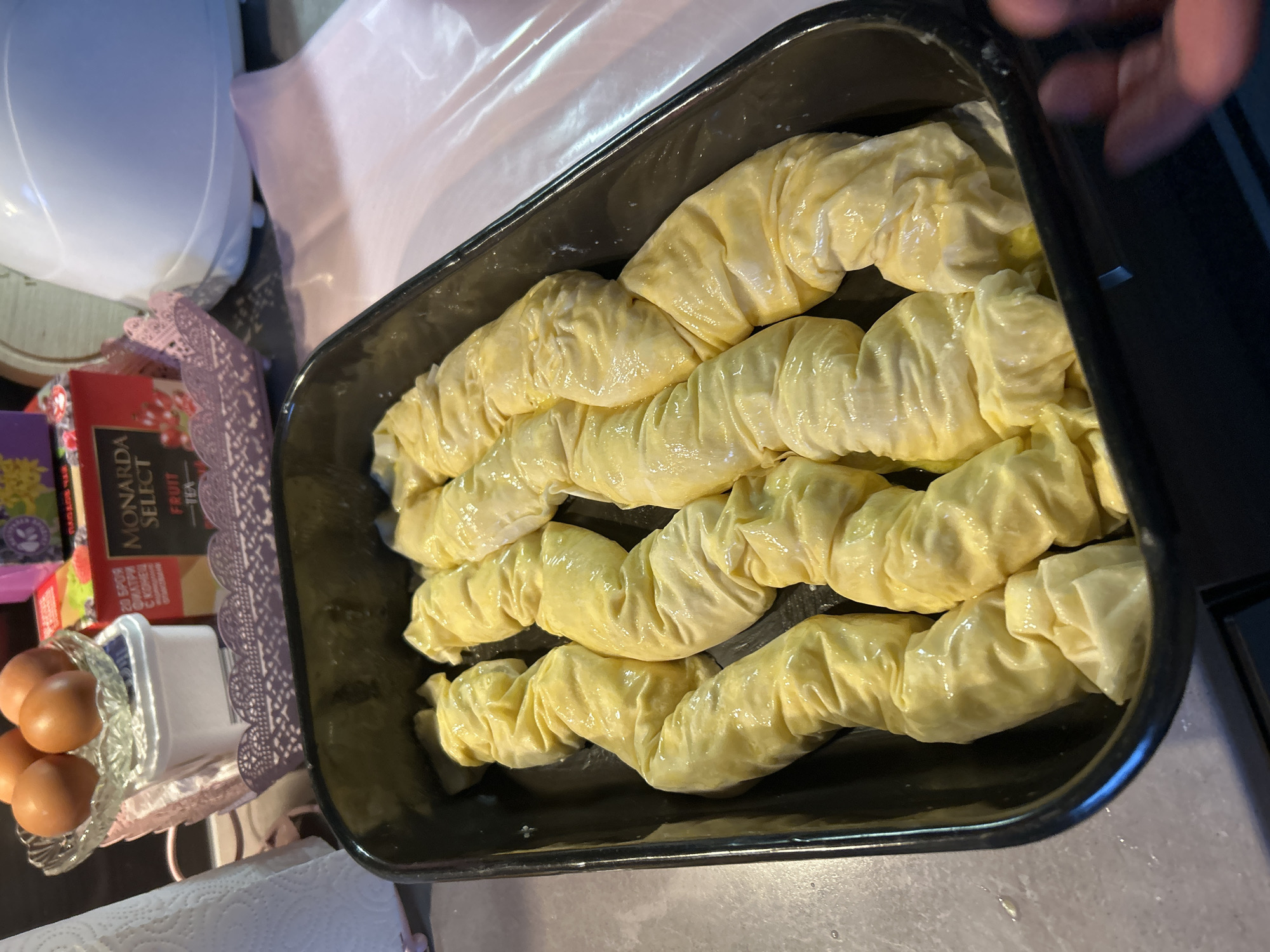 A top-down view captures a black rectangular baking pan filled with four rows of pale yellow, rolled pastries, likely pre-baked or partially baked. The pastries appear glossy, suggesting a coating of egg wash or oil. The pan is situated on a kitchen countertop, with a portion of a white kitchen appliance visible on the left. To the left of the pan are a container of eggs and a box of tea, arranged on a surface covered with a pink doily. The corner of the pan is being held by a human hand. The setting is indoors, potentially in a residential kitchen. Krumovo, Bulgaria is not suggested by anything in the scene.
