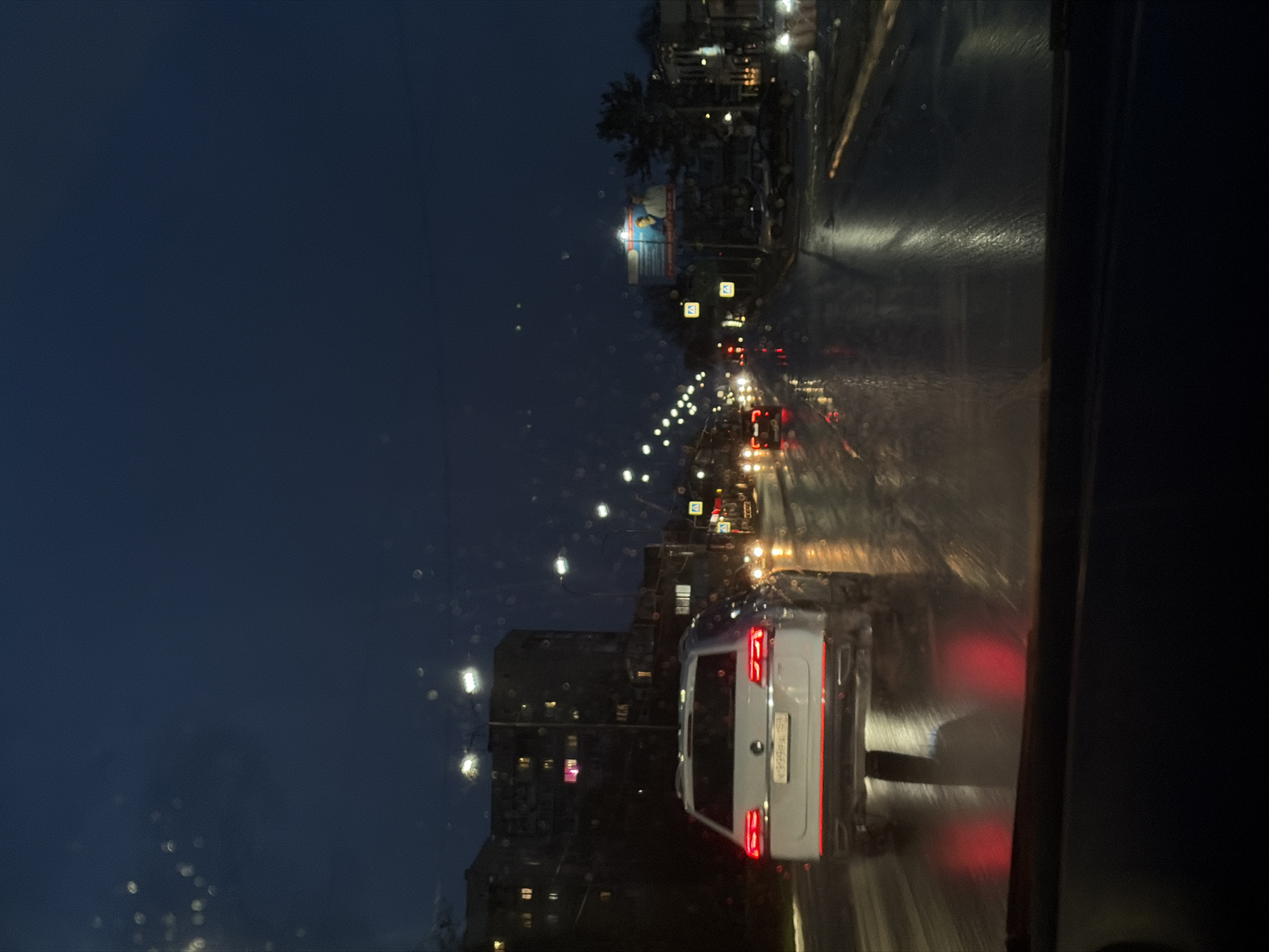 The image captures a nighttime urban street scene from the perspective of a vehicle looking forward through a wet window. The road, slick with rain, reflects the numerous bright streetlights and car lights stretching into the distance. In the right foreground, the rear of a white minivan or small SUV is visible, its red taillights glowing prominently. The license plate on this vehicle reads "Х999АА 59," indicating a Russian registration from the Perm Krai region (which includes Chaikovsky).
To the left of the busy street, multi-story buildings line the thoroughfare, some showing illuminated windows. The sky above is a dark, deep blue, consistent with late evening or night. Water droplets and streaks on the windshield or window create a soft, diffused halo effect around the light sources. The scene suggests a busy city road at night, during or immediately after rainfall, creating a reflective and atmospheric setting in Chaikovsky, Russia.