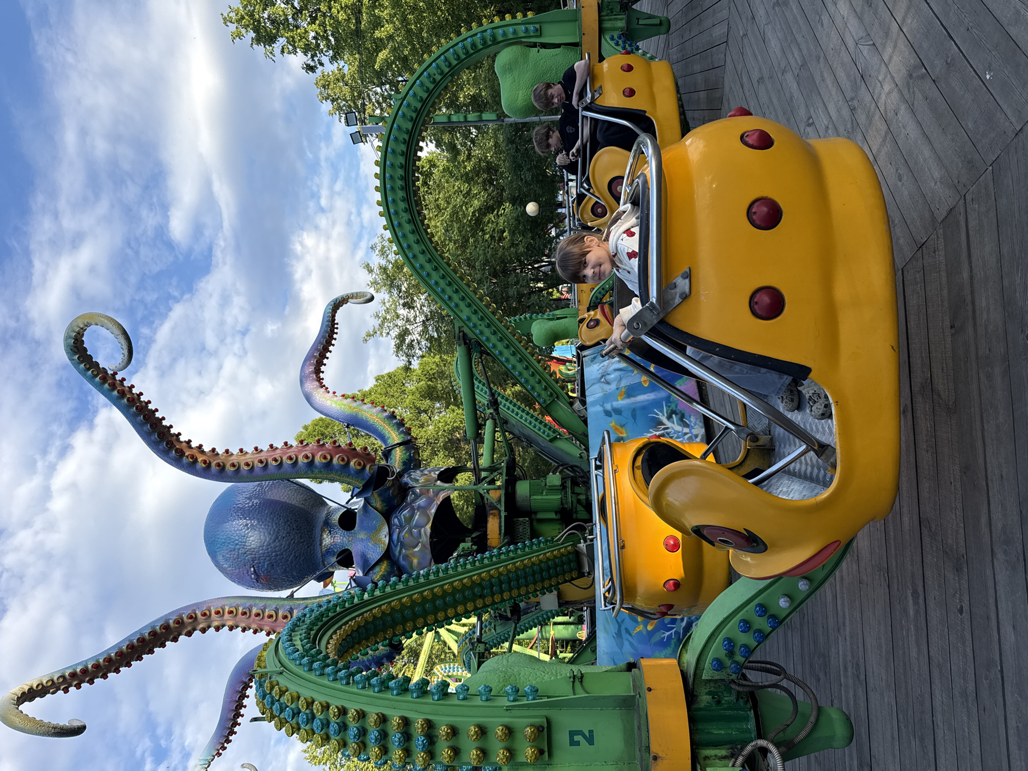 An outdoor, vertically oriented eye-level shot captures a vibrant octopus-themed amusement park ride in Заволжье, Russia. In the foreground, a young girl with brown hair, wearing a white and red patterned jacket, sits in a bright yellow ride car, looking directly at the camera with a slight smile. Her hands are on the safety bar in front of her. Behind her, two boys are visible in another yellow car, looking forward.
The central feature of the image is the large, colorful octopus ride. Its massive blue head is adorned with purple and blue hues, and multiple green and purple tentacles, detailed with rows of colorful suckers, curl upwards towards a bright blue sky with scattered white clouds. The ride cars are attached to a large green support structure, which is also decorated with rows of yellow and blue light fixtures, suggesting it might illuminate at night.
The ground beneath the ride is a wooden plank platform. Lush green trees are visible in the background, framing the top portion of the image. The time of day appears to be sunny daylight. The atmosphere suggests a fun and lively event at an amusement park. The number "2" is visible on a green metal support beam of the ride structure.