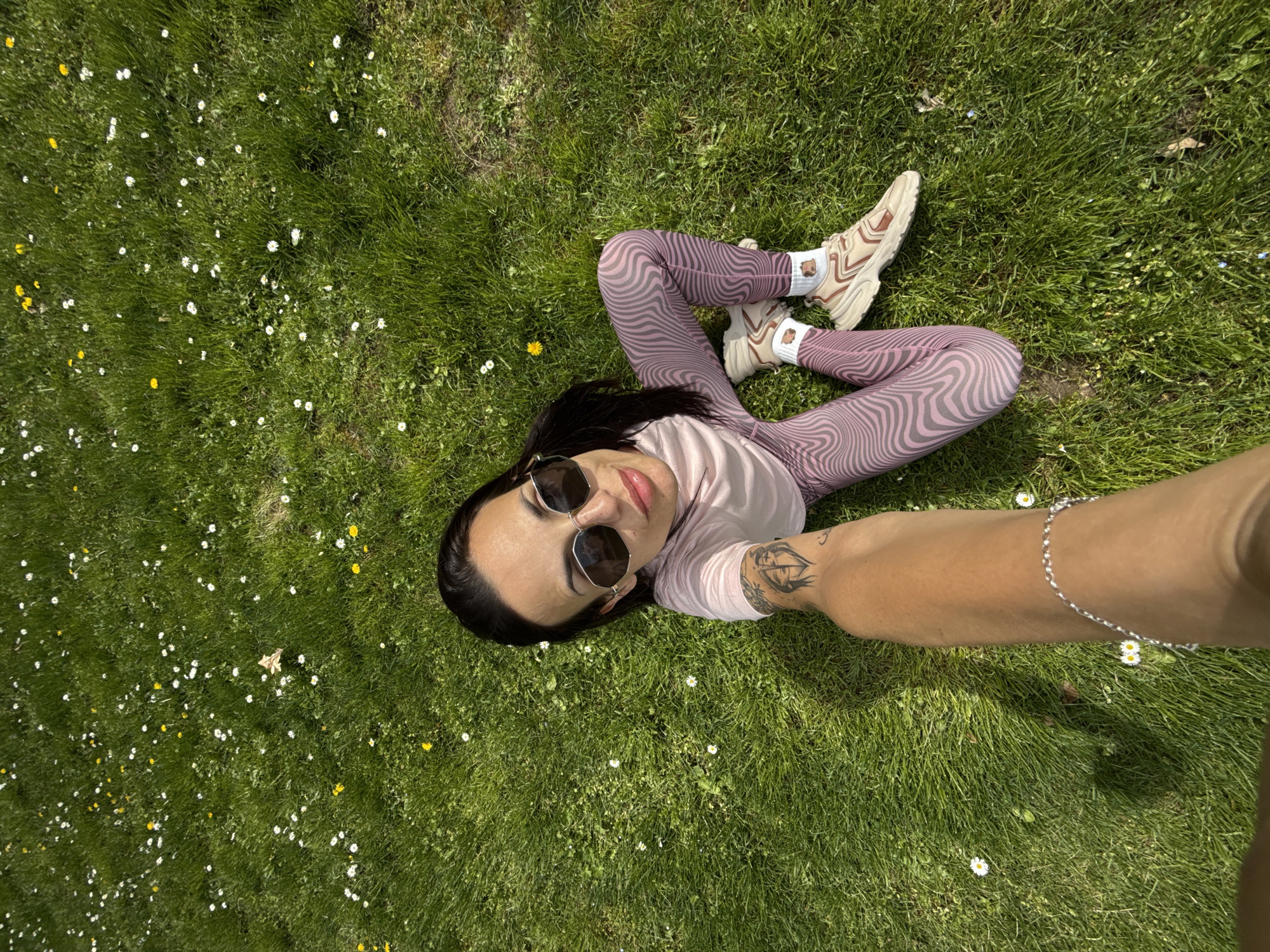 A woman is lying on a grassy field. The grass has white and yellow flowers. She is wearing leggings and a light-colored t-shirt, sunglasses, and sneakers. Her left arm is extended holding the camera for the photo. Her left arm has a tattoo. The location is Pleven, Bulgaria.