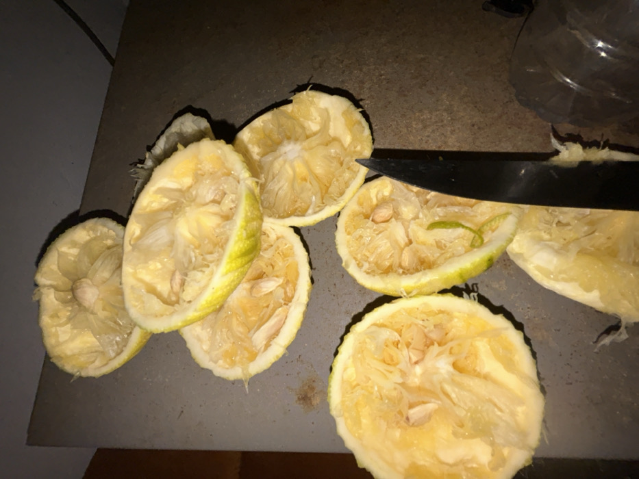 The image displays a close-up, overhead view of several halved and squeezed citrus fruits, likely lemons, resting on a dark, flat surface, possibly a countertop or cutting board. The fruits are mostly depleted of their juice, showing the fibrous pulp and numerous seeds within their rinds. Their inner flesh is a pale yellow, while the rinds exhibit varying shades of yellow and a touch of light green.
A black-bladed knife is partially visible in the upper right portion of the frame, resting among the squeezed fruit halves, suggesting it was used to cut them. In the upper right background, a blurred, translucent object that appears to be a plastic bottle or container can be faintly seen.
The lighting in the image is somewhat dim, creating distinct shadows around and beneath the fruits, particularly noticeable on the left side. The overall scene suggests the aftermath of juicing, possibly for cooking or making a beverage. There is no visible text in the image.