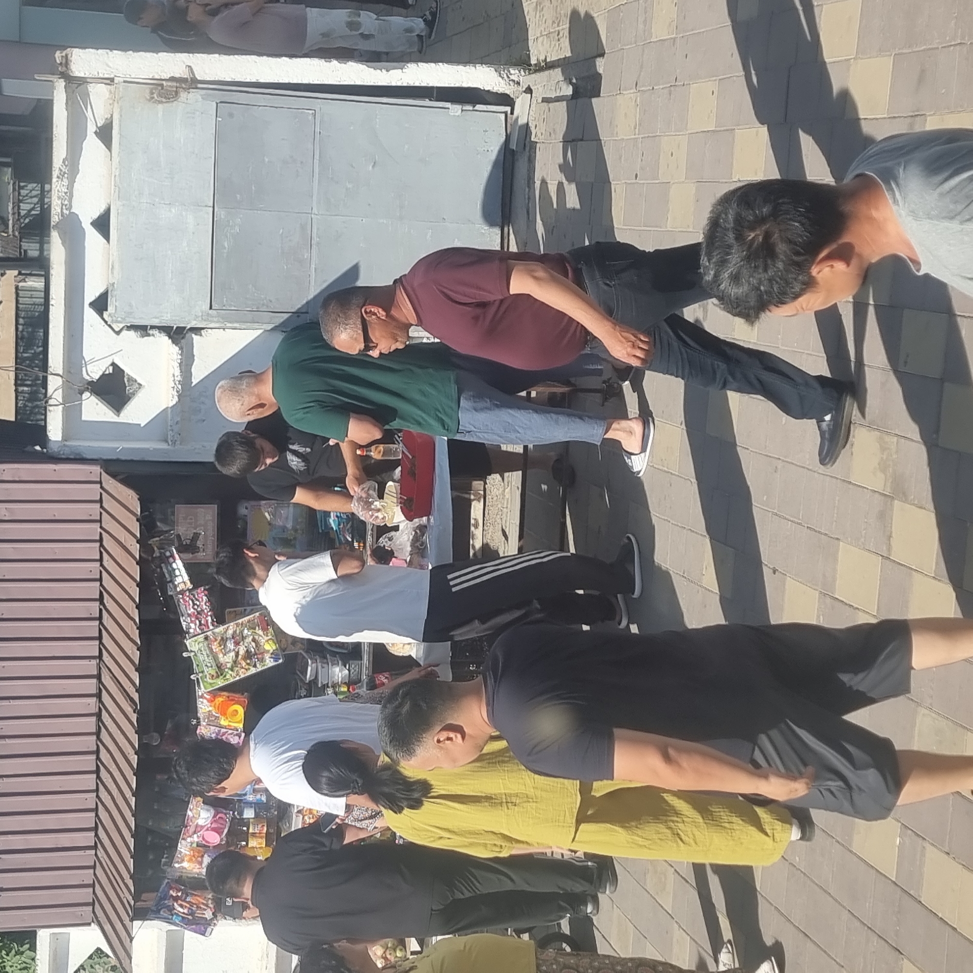 A group of people are gathered around a small outdoor stall selling various items, likely toys and snacks, under a brown awning. The stall is set up in a paved outdoor area with a checkered pattern of light and dark square tiles. Behind the stall is a building with a corrugated metal roof and a light grey metal gate.
In the foreground, from left to right, a person in a yellow outfit stands with their back to the camera. To their right, a man in a black t-shirt and grey shorts is walking away from the viewer. Further right, a man in a maroon t-shirt and dark jeans walks past, looking towards the left. Another man in a grey t-shirt is partially visible in the far right foreground, his head turned away from the camera.
Near the stall, several people are interacting. A man in a green t-shirt and blue pants is handing something from a red container to a man in a black t-shirt. Another man in a white t-shirt stands nearby, looking towards the stall. A woman in a black t-shirt and patterned pants is on the far left, mostly obscured.
The scene appears to be a market or street-side vendor location. The overall impression is a busy, casual street scene. This is a scene consistent with the general street vending environments that can be found in the city of Turkistan, Kazakhstan, particularly in areas with pedestrian traffic and smaller commercial establishments.