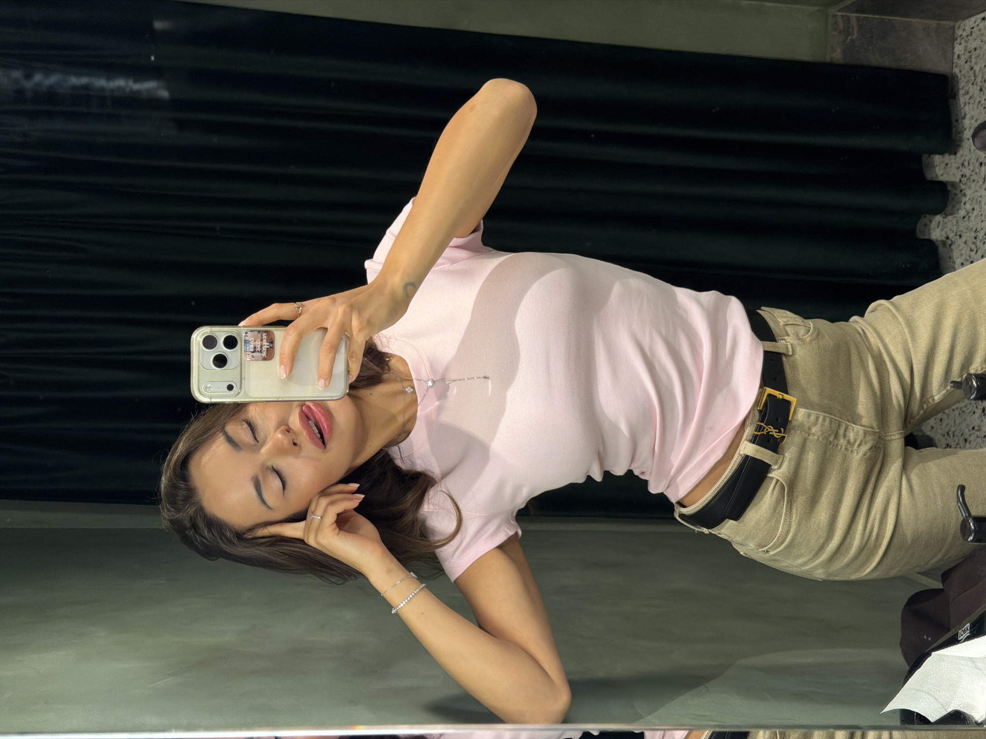 A woman in a pink t-shirt and tan pants is taking a selfie in a fitting room. She has her eyes closed, tongue sticking out, and her left hand touching her forehead. She is wearing a black belt with a gold YSL buckle, a silver bracelet, and a necklace. The background appears to be a dark green curtain. The location is a fitting room in Saint Petersburg, Russia.