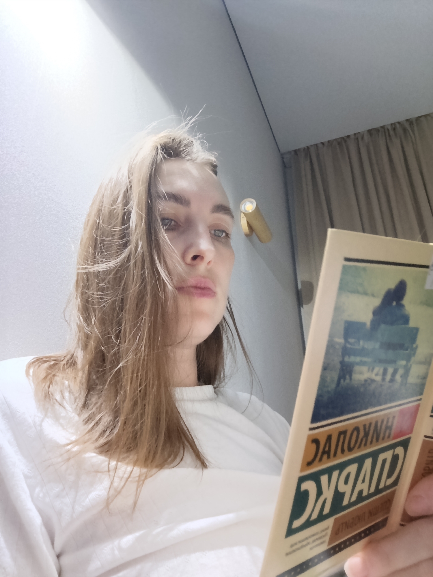 A woman is holding a book in front of her face, indoors, seemingly reading. The book cover has text in Russian and depicts an image of two people sitting on a park bench. A gold-colored light fixture is visible on the wall behind her. The woman is wearing a white shirt. Based on the book's cover, it is likely that the photograph was taken in the city of Chaikovsky, Russia.