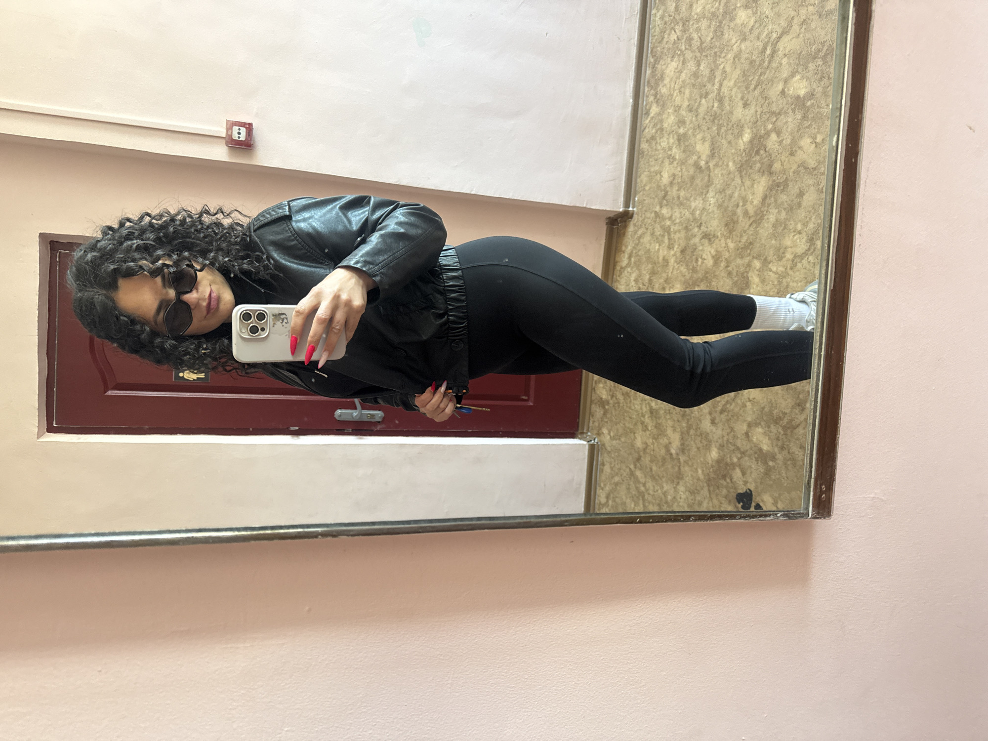 A young person is captured in a mirror selfie, standing upright and posing indoors in Chaikovsky, Russia. The individual has dark, curly hair and is wearing dark sunglasses, a black leather-style jacket, black leggings, white socks, and light-colored sneakers.
The person is holding a light-colored smartphone with a visible multi-lens camera array in their right hand to take the mirror selfie. Their right hand features long, pointed, bright red fingernails. Their left hand holds a small bunch of keys with a blue and yellow/orange tag.
The reflection in the mirror reveals the setting: a light pink/peach colored wall, a section of textured light brown/beige flooring, and a dark red/maroon door. Mounted on the pink wall, above the person's reflection, is a red fire alarm box with a white button. A small pictogram sign, depicting male and female figures, is visible on the red door, suggesting it leads to a restroom. Below the door, a horizontal metallic bar or rail is also reflected. The mirror itself has a thin, possibly metallic frame.