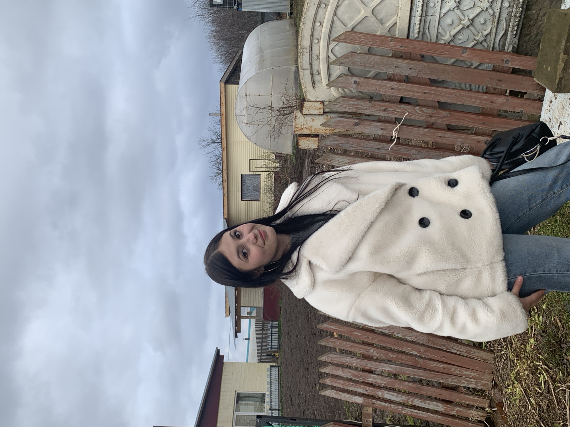 The main subject of the image is a young woman, seen from the waist up, positioned outdoors in what appears to be a rural or semi-rural setting in Фоки, Russia. She has long, dark hair and is looking directly at the camera with a gentle smile. She is wearing a cream-colored, fuzzy winter coat with prominent dark buttons and blue jeans. A black handbag with a silver zipper rests by her right side.
She is standing or leaning in front of a rustic, weathered wooden fence with horizontal slats. Beyond the fence, the background reveals various structures. To the left, a large, ornate, circular white object with decorative patterns is visible, possibly a well cover or a large garden feature. Next to it, a translucent, tunnel-shaped greenhouse stands. Further in the background, several single-story houses with light-colored siding (yellow or cream) and dark roofs are visible. One house features a small window with a white curtain. The ground in the foreground and between the structures consists of sparse dry grass and patches of dirt, suggesting late autumn or early spring. Bare trees and bushes are scattered throughout the scene. The sky is completely overcast with light gray clouds, indicating a cloudy day. No visible text is present in the image.