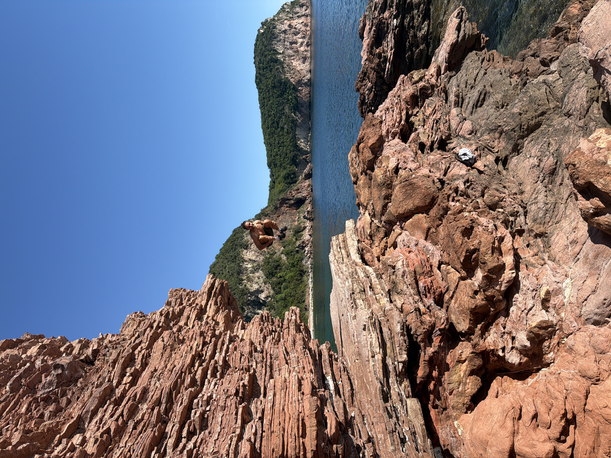 The image depicts a coastal scene in Sveti Stefan, Montenegro. A person is captured mid-jump, seemingly diving from a rocky cliff into the water below. The foreground features red and brown rocks, displaying a layered and textured surface. In the background, a forested hill extends into the clear blue water, with a cloudless blue sky above. The overall composition is a vibrant landscape showcasing a daredevil cliff jump.