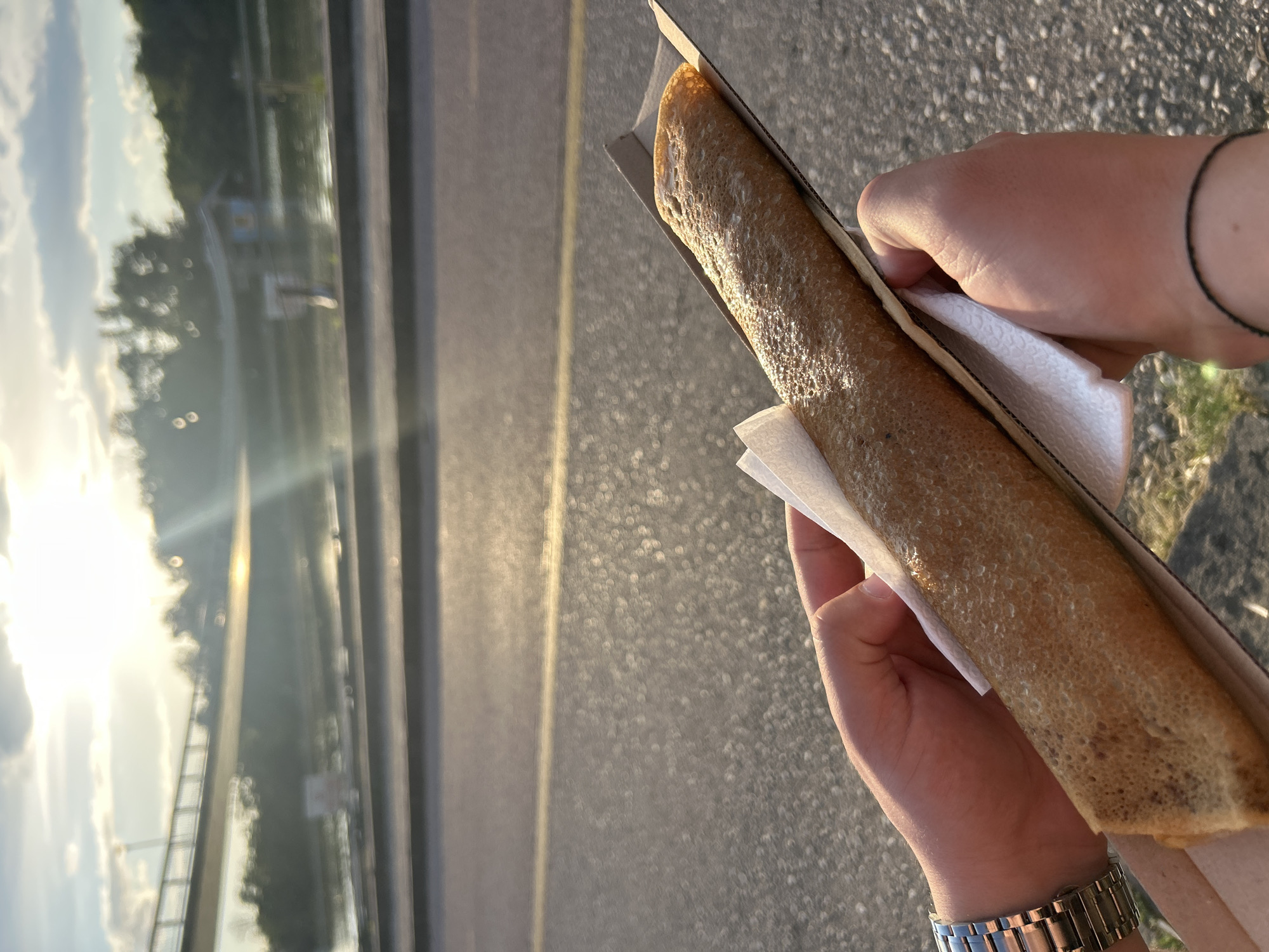 A person holds a long, rolled crepe in a cardboard container. The crepe is golden brown with a slightly textured surface. The person is holding it with both hands, using a white napkin to protect their hands. A silver watch is visible on one wrist, and a simple black bracelet on the other. The background is a slightly blurred asphalt surface with a yellow line. In the distance, there is a view of a river or body of water with trees on the opposite bank, illuminated by the setting sun. This appears to be in Plovdiv, Bulgaria near the Maritza river.