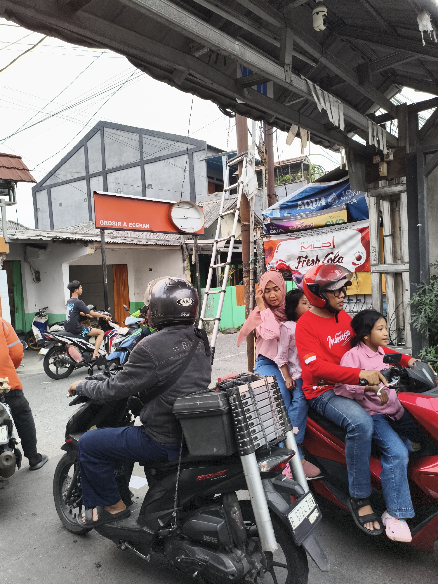 The image shows a street scene in Depok, Indonesia. Motorbikes are the primary mode of transportation depicted. One motorbike carries a family of four: a man wearing a red shirt and helmet drives the bike, while a woman in a pink hijab and two young girls sit behind him. A ladder is strapped to the back of the motorbike. Another motorbike is directly in front of them, carrying a man wearing a black helmet. Several other motorbikes are further down the street. A building with an orange sign that says "GROSIR & ECERAN" is visible. Above the family on the motorbike is a sign advertising a soft drink. Overhead, wiring and parts of a building structure are visible.