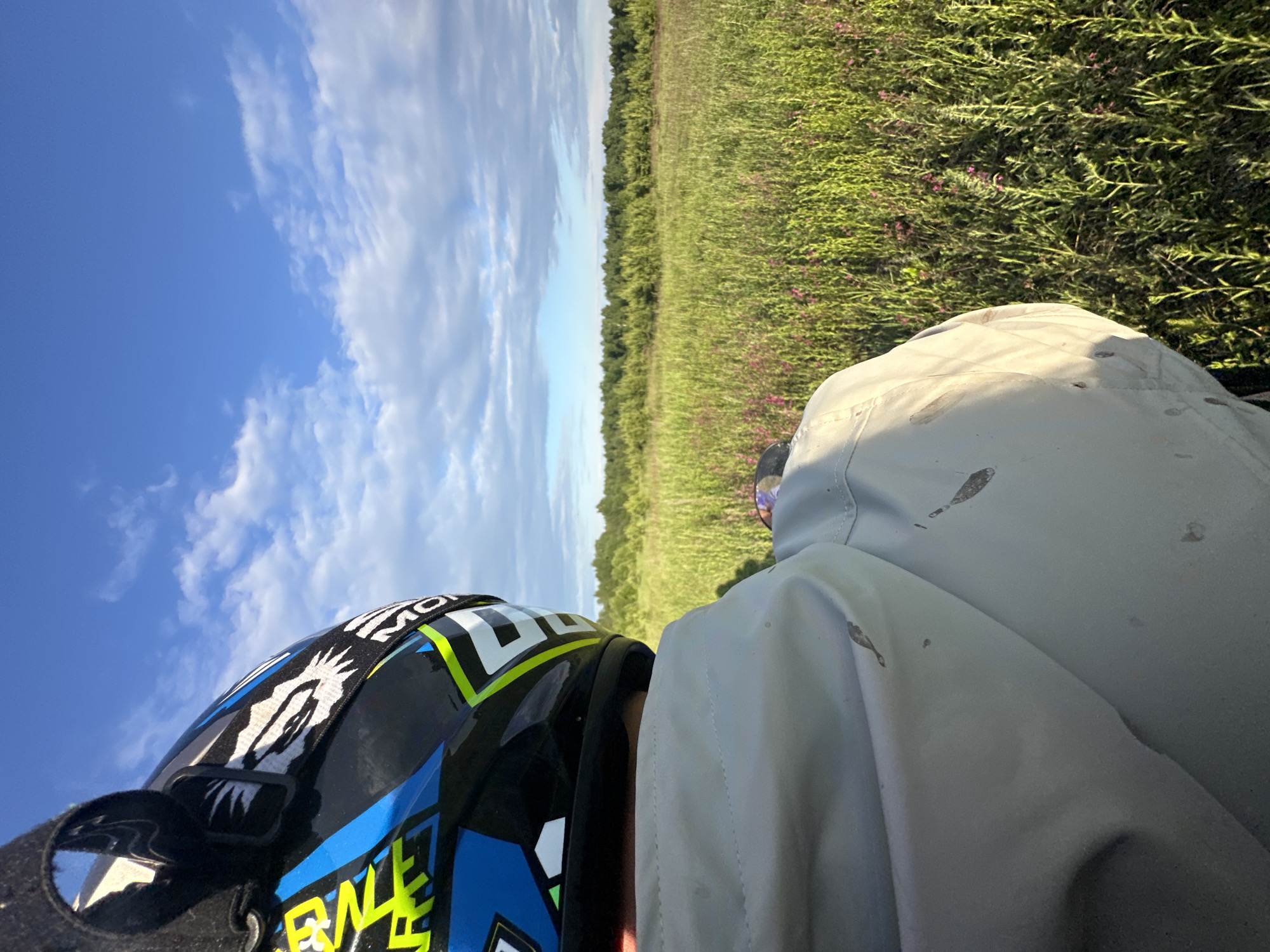 The image depicts a person viewed from behind, situated in an open, natural environment identified as Kharnavy, Russia. The individual wears a blue, black, neon yellow, and white motocross-style helmet with prominent graphic designs, including what appears to be a stylized creature or animal head on the side and the partial word "MOTO" visible on the strap. A small, round, reflective object is attached to the goggle strap on the left side of the helmet. The person is also wearing a light gray or off-white jacket, which shows several visible dark smudges or stains on the back and shoulder areas. The subject is oriented to observe a vast field of tall, vibrant green vegetation that fills the midground, interspersed with clusters of small pink or purple flowers. A dense line of green deciduous trees forms the distant horizon, separating the field from a clear blue sky dotted with scattered white cumulus clouds. The lighting suggests natural daylight, casting subtle shadows and highlighting textures.