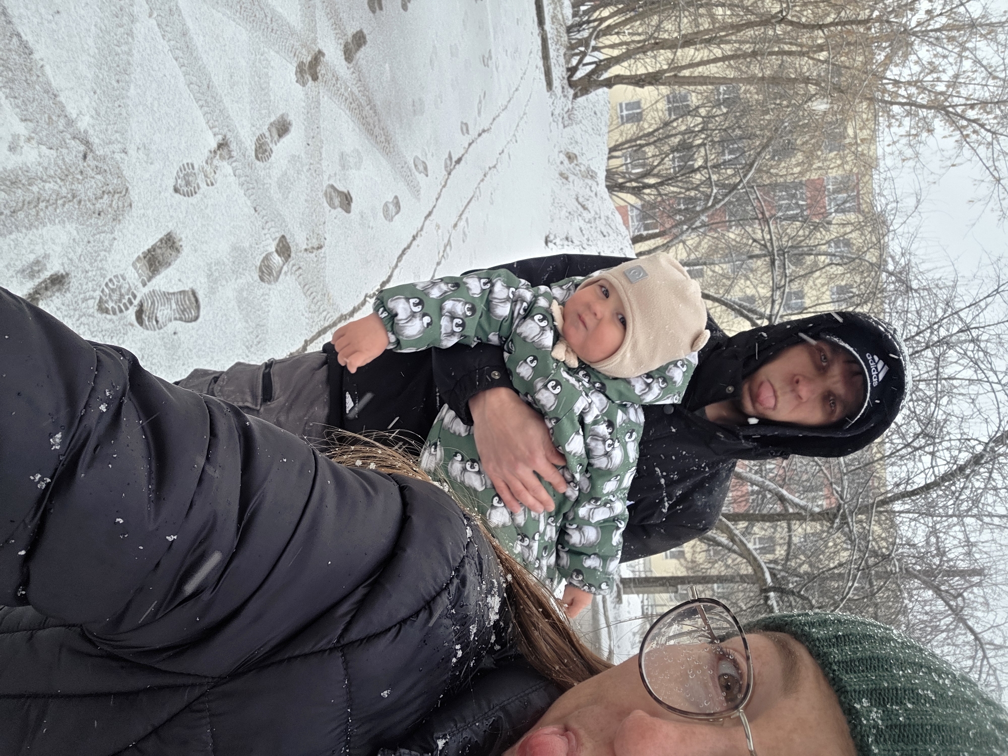 The image depicts a snowy scene, likely in the city of Chaikovsky, Russia, as indicated by metadata not included in the request. A family, consisting of two adults and a young child, appears to be outside. The ground is covered in snow, with footprints visible on a path. The backdrop includes a building and bare trees, suggesting winter.
One adult is holding the child, who is wearing a green and white penguin-patterned snowsuit and a beige hat. This adult is wearing a black hooded jacket. The other adult is taking the picture, as part of their face and glasses are visible in the lower portion of the frame. They are wearing a green hat and a black jacket covered in snowflakes. All three individuals have their tongues stuck out.