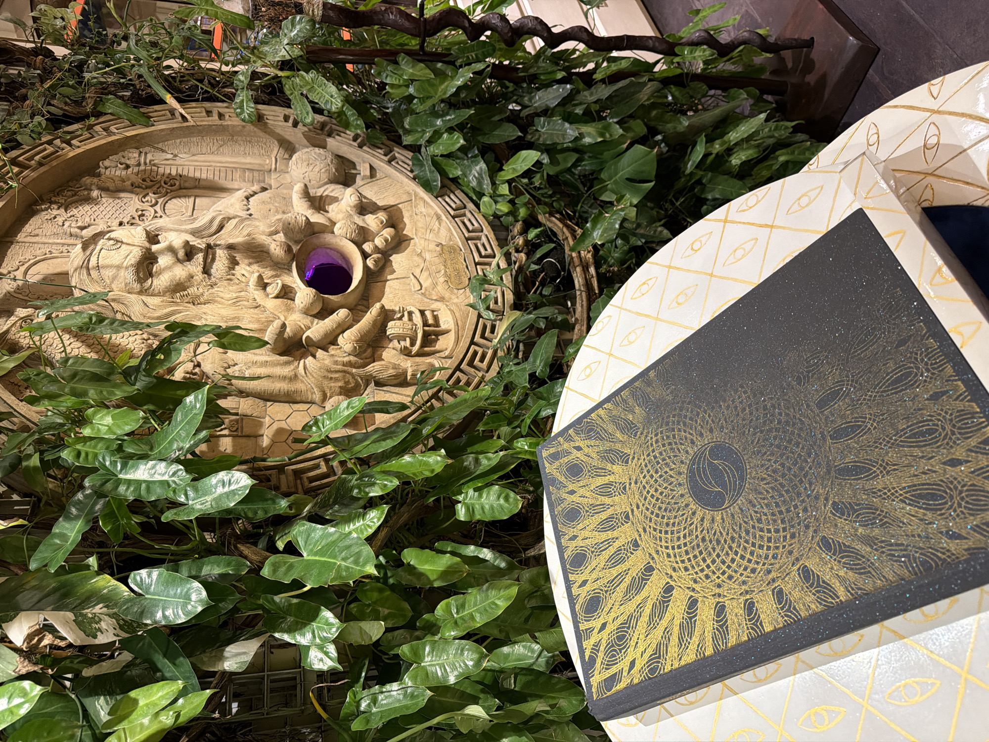 In Ubud District, Indonesia, a round, intricately carved wooden relief depicts a mystical figure with a long beard and flowing hair. The figure's hands are outstretched, and in the center of its chest is a circular, purple void. This relief is set against a lush backdrop of green foliage, with large, heart-shaped leaves cascading down. In the foreground, a table with a white and gold patterned surface holds a dark-colored book. The book is adorned with a golden, intricate design resembling a sunburst or peacock feathers, and it appears to have a slightly textured, glittering cover. The overall scene evokes a sense of enchantment and mystery, fitting for a location like Ubud, known for its spiritual and artistic atmosphere.