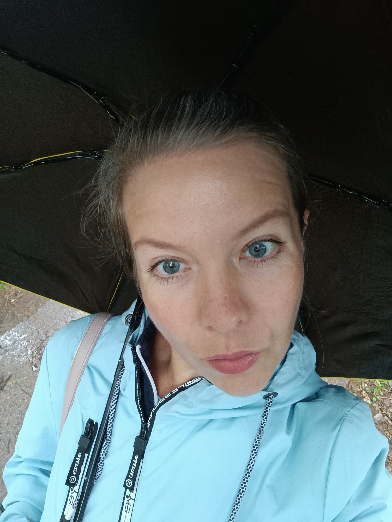 A woman is holding a black umbrella over her head, seemingly outdoors. She is wearing a light blue hooded jacket with a white and black patterned drawstring. The jacket has a zipper and branding visible on the zipper pulls. She has light skin, blue eyes, and her hair is pulled back. The background shows a glimpse of a wet, possibly paved surface, and some greenery. The photo appears to be taken from a slightly elevated angle, looking down on the subject. The location is Chaikovsky, Russia.
