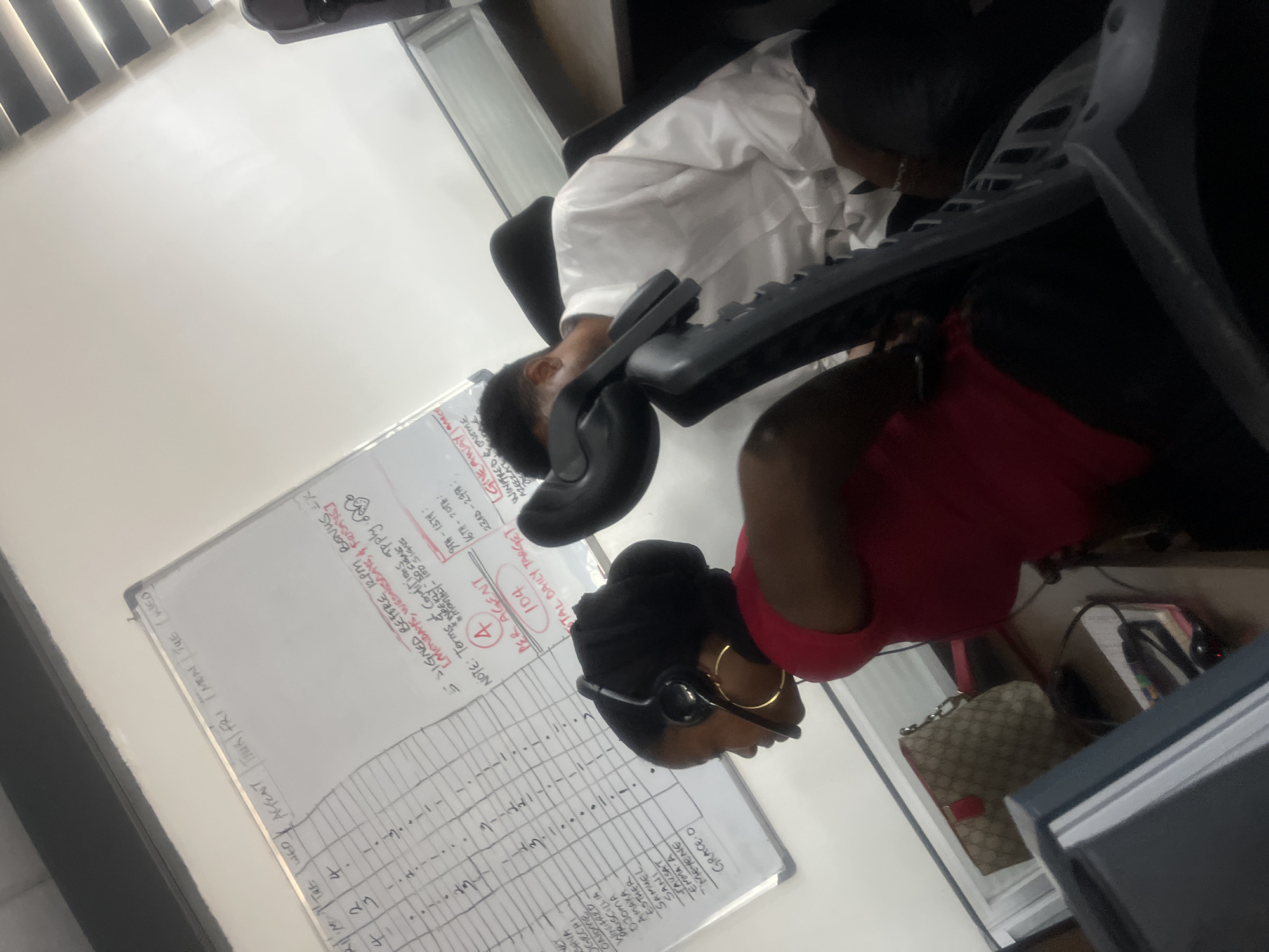This media file captures an indoor office scene in Karum, Nigeria, featuring two individuals engaged in work-related activities. The image is oriented sideways, but depicts an office environment with white walls, office furniture, and a prominent whiteboard.
One person, wearing a white long-sleeved shirt, is seated in an office chair, leaning forward with their head resting on what appears to be a computer keyboard or desk. They seem to be either resting or asleep. Adjacent to them, another individual is visible from the side, wearing a red sleeveless top and a dark head covering. This person is wearing a black headset with a microphone and large gold hoop earrings, suggesting they are on a call or actively working at a computer station, though the monitor is mostly out of frame. A patterned handbag, possibly a Gucci design, is visible on the floor or a shelf near this person.
A large whiteboard is prominently displayed in the background, filled with handwritten text and a grid, indicative of tracking work performance or targets. Visible text includes names like "ONOME", "WINIFRED", "AMAKA", "ESTHER", and "GRACE O". There are also sections detailing work incentives and targets: "SIGNED BEFORE 12PM, BONUS K", "MONDAYS, WEDNESDAYS, & FRIDAYS", "NOTE: Terms ContiTpply", "PER AGENT 104", "TOTAL DAILY TARGET 100 SIGNS". Specific date ranges like "9TH-13H", "16TH-20TH", and "23RD-27" are also present, alongside a "GIVEAWAY" section mentioning "WINPFRED & ONOME" and "AMEEZAT". The general atmosphere suggests a busy call center or customer service environment during what appears to be working hours.