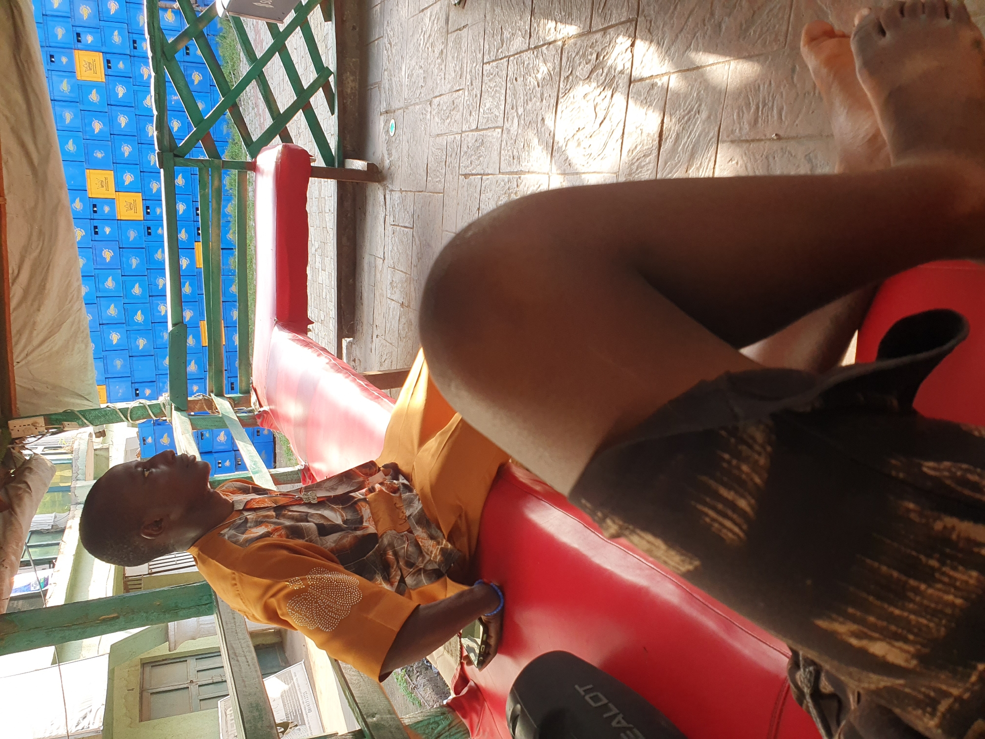 The image depicts two individuals in an outdoor setting, likely a rest stop or a shaded outdoor seating area in Lokoja, Nigeria. The foreground is dominated by the legs of one person, partially obscured by a dark, patterned fabric, sitting on a red cushioned bench. The second person, a male, is visible in the mid-ground, seated on the same bench. He is wearing an orange traditional outfit adorned with intricate beadwork on the left shoulder and chest. His gaze is directed to his right, away from the camera.
The environment suggests a warm climate. Behind the individuals, a wall of stacked blue crates, possibly for beverages, is visible. A green wooden lattice structure separates the seating area from the crates. The ground is paved with textured concrete tiles, with sunlight casting distinct shadows, indicating it is daytime. A black object, possibly a speaker or bag, is placed on the red bench in the foreground. A small mobile phone is visible near the right hand of the seated male. The overall atmosphere is casual and relaxed.