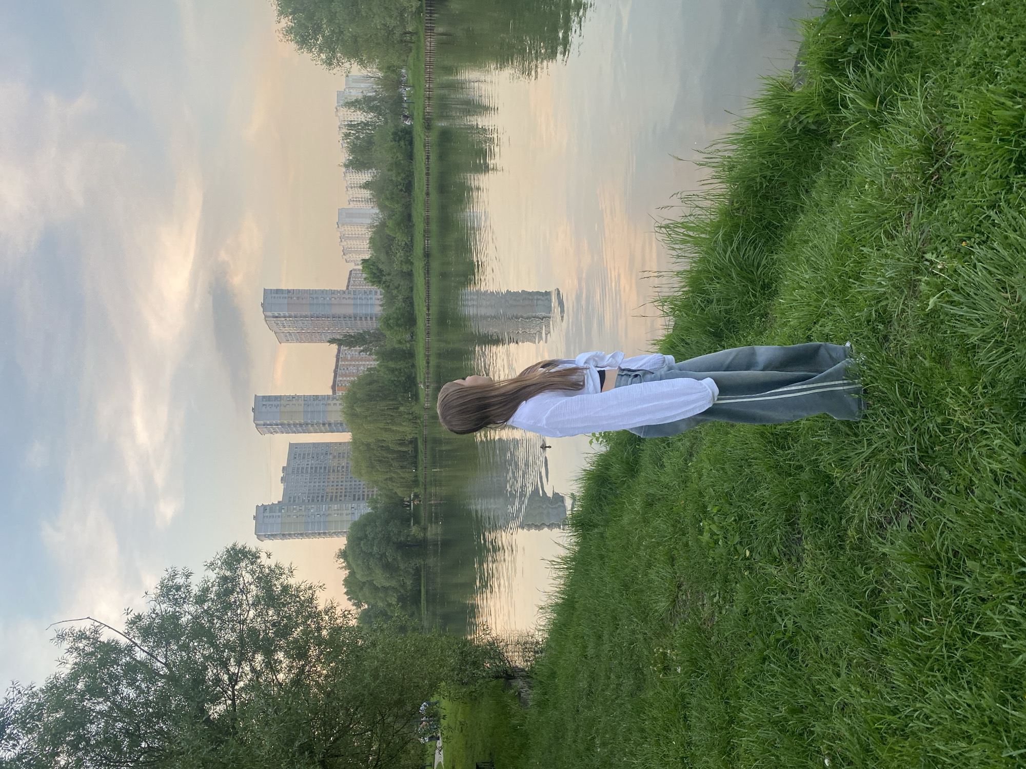 A woman lies on a grassy bank beside a body of water, possibly a lake or river, in a park setting. The location is Moscow, Russia, as indicated by the architecture of the buildings visible in the background.
The woman has long brown hair and is wearing a white shirt and gray pants. She is lying on her back, with her head facing the water and her gaze directed upwards.
The surrounding environment includes lush green grass, trees, and a body of water that reflects the surrounding sky and buildings. The buildings are high-rise apartments or office buildings, typical of urban areas. The sky is overcast with light clouds.