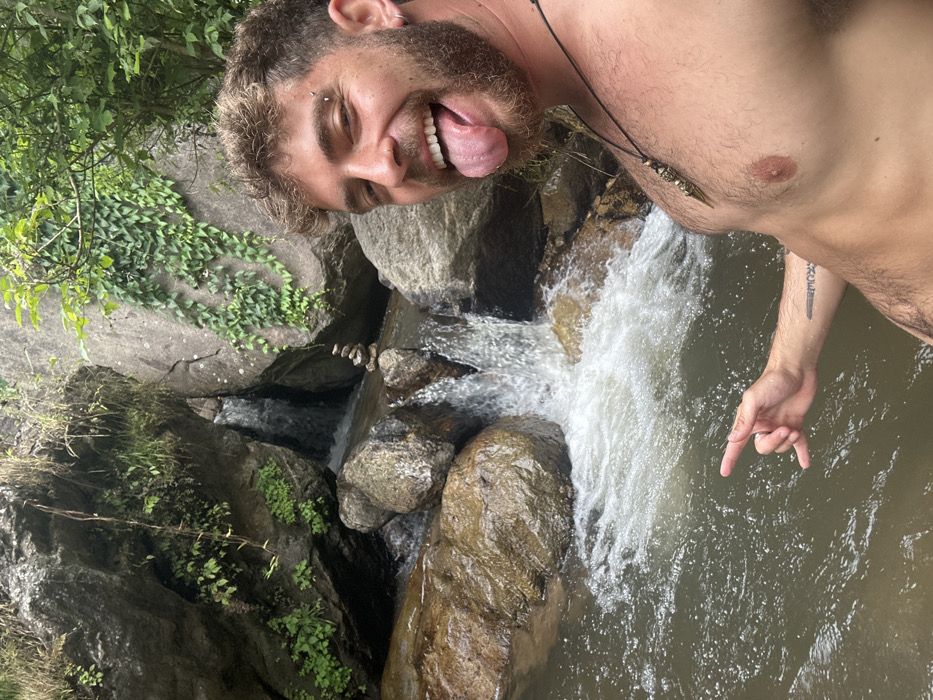 The image displays an adult male posing in a natural environment identified as Ban Mai Saha Sampan, Thailand. The man, positioned on the right side of the frame, is shirtless, has a beard, curly brown hair, a nose ring, an eyebrow piercing, and an earring. He wears a dark necklace with a metallic, ornate pendant. His left hand, extended towards the left and partially submerged in water, forms a 'shaka' gesture. He is smiling broadly with his tongue extended and looking directly at the camera. The background features a stream with water flowing over and around large rocks, forming a small waterfall or series of cascades. A small cairn (stack of balanced stones) is visible on a rock in the center of the stream. Lush green vegetation, including vines and smaller plants, covers the rocks and banks, indicating a verdant, rocky, and watery landscape.