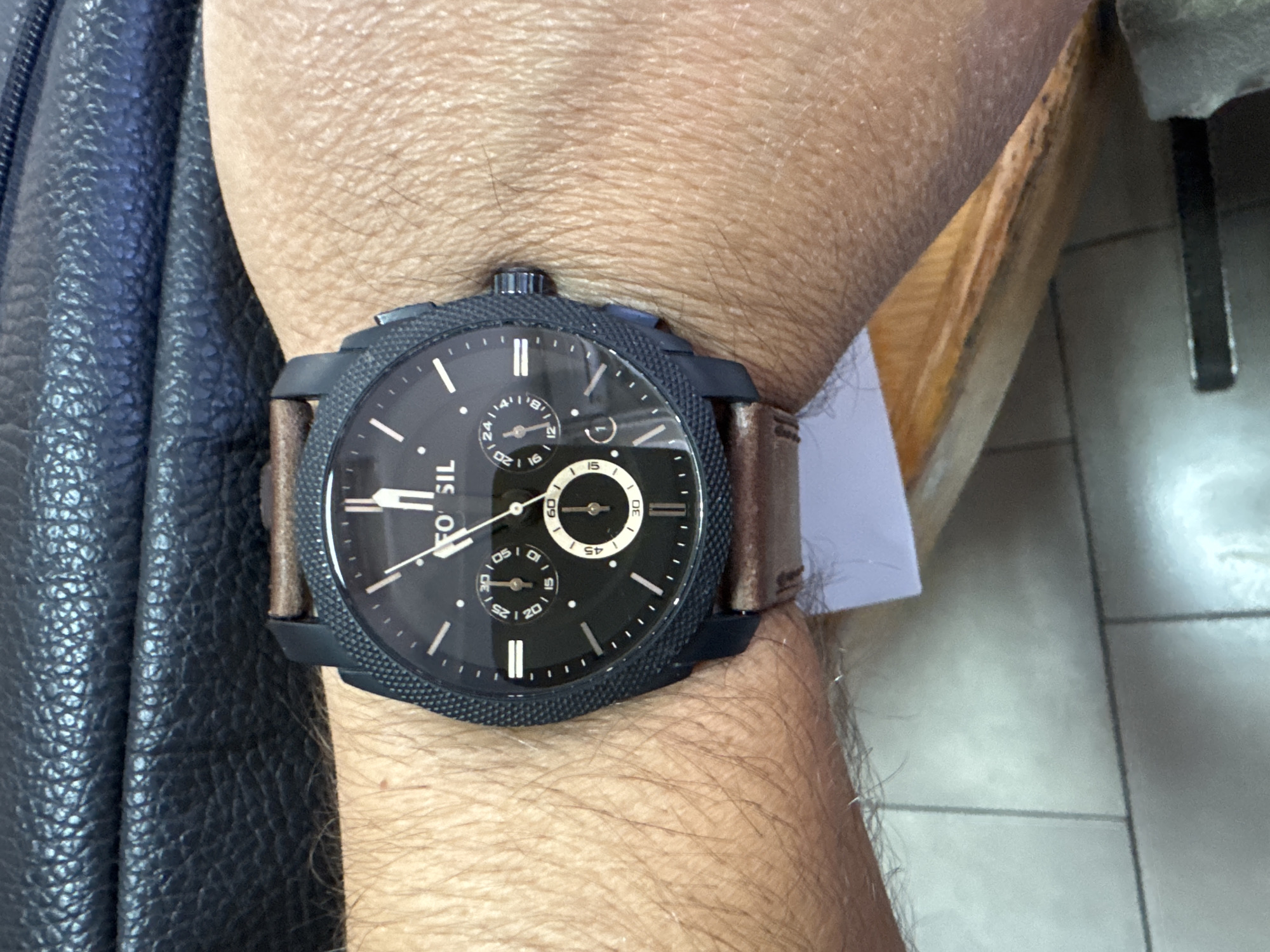 A close-up shot depicts a person's wrist with a Fossil brand chronograph wristwatch. The watch features a dark-colored textured bezel, a black dial with white markers and hands, and brown leather straps. The watch face includes subdials and a date display. The person is wearing a black leather jacket, which is visible on the left side of the image. The wrist is surrounded by skin with visible hair. The background shows a section of a light-colored surface, potentially a wooden chair or table, and a glimpse of light-colored tiled flooring.