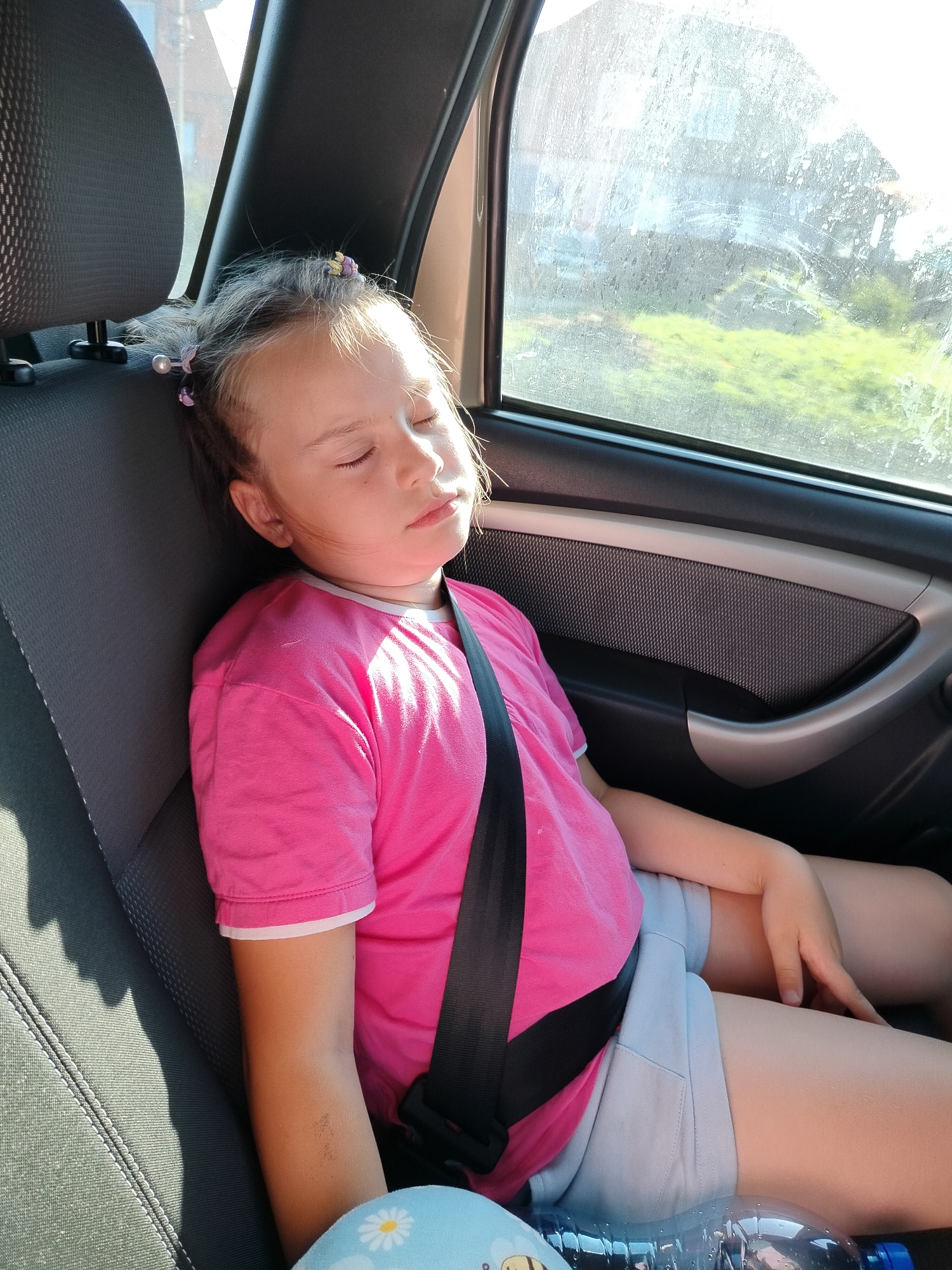A young girl with blonde hair styled in pigtails with colorful clips is sleeping in the back seat of a car. She is wearing a bright pink t-shirt with white trim and light blue shorts. A black seatbelt is fastened across her chest. The girl's eyes are closed, and her head is resting against the car's headrest. Sunlight streams in from the window, illuminating her face and casting shadows on the dark gray car seat. Outside the window, green foliage and a building are visible through the slightly dirty glass, suggesting a daytime scene in a rural or suburban area. A water bottle lies on the seat in front of her. The image appears to capture a moment of rest during a car journey.