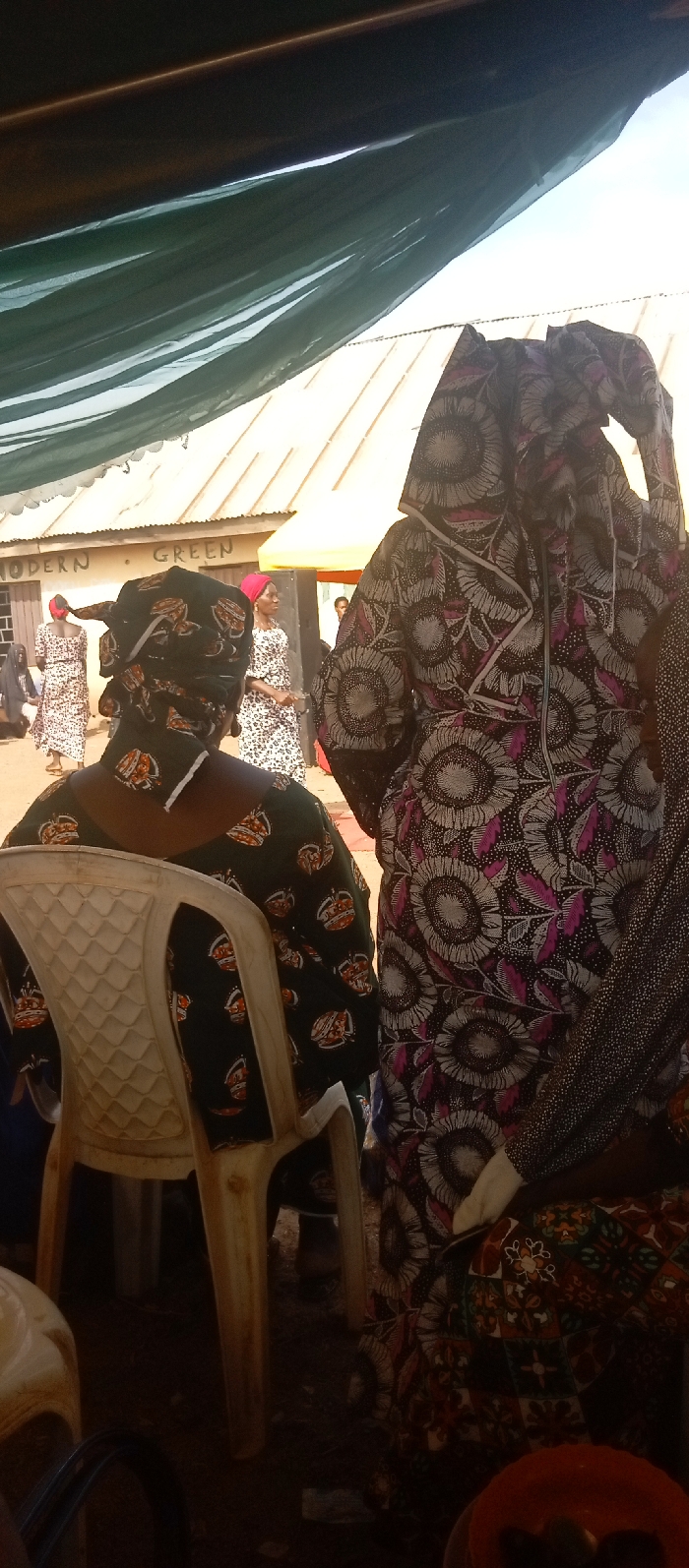 The image captures an outdoor gathering in Kasuwan Magani, Nigeria, viewed from beneath a dark green fabric canopy. In the immediate foreground, a person is seated with their back to the viewer on a light-colored plastic chair, wearing a dark green garment patterned with orange and brown crown-like designs, and a matching headwrap. To their right, another individual is partially visible, dressed in a dark fabric featuring large grey and white sunflower-like patterns with pink and magenta leaves.
In the midground, multiple individuals are present in an open, unpaved area. Two women are visible, both wearing similar light-colored, spotted dresses; one has a distinct red headwrap. A light-colored building with a corrugated metal roof, marked with the words "MODERN GREEN," stands in the background. A dark, rectangular object, possibly equipment, is positioned near this building. The scene is illuminated by natural daylight, casting shadows and indicating an outdoor environment with an unpaved ground surface.