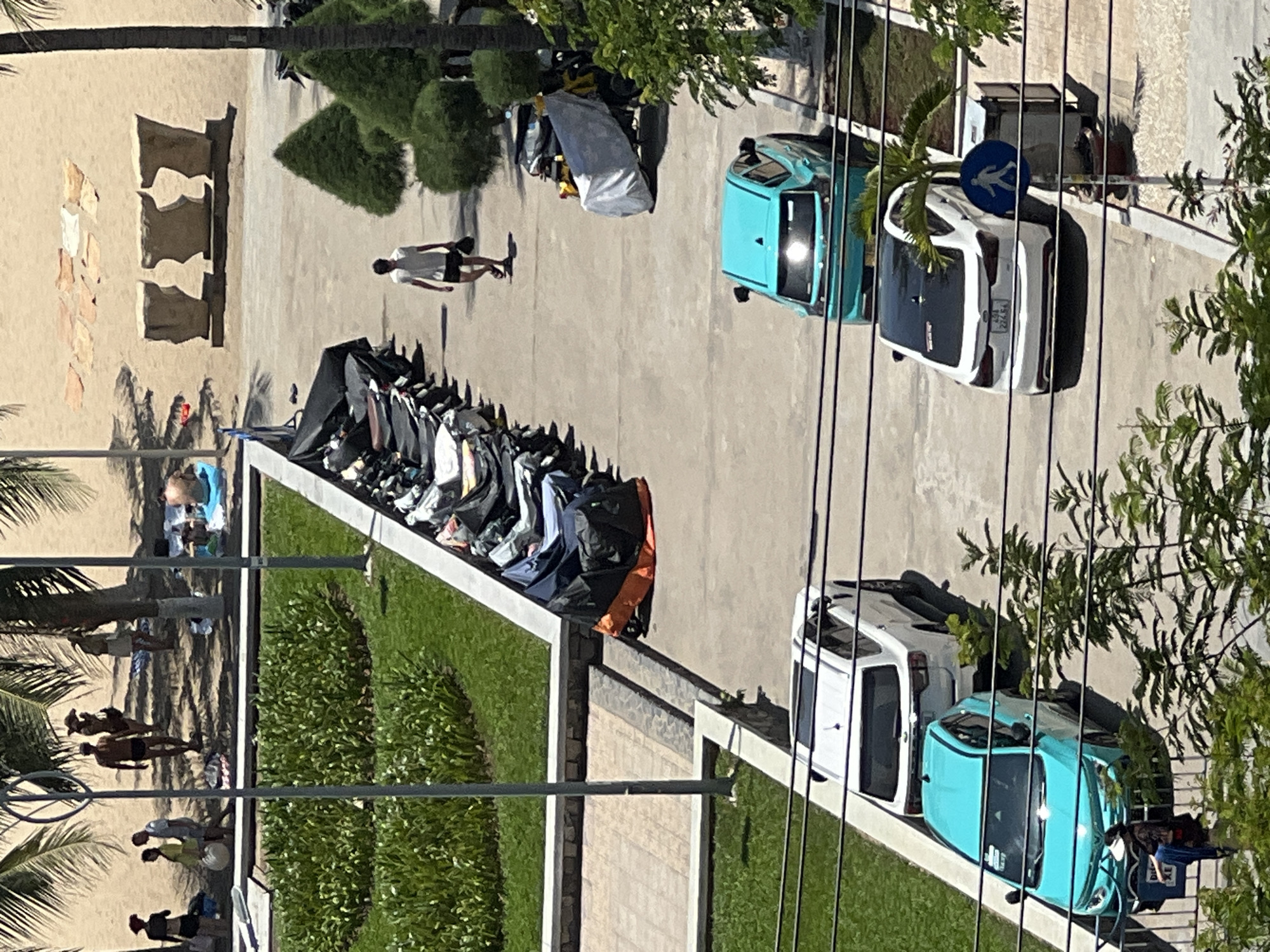 The image captures a scene from an elevated perspective, looking down onto a street adjacent to a beach area in Nha Trang, Vietnam. Palm trees and manicured green spaces flank the paved pathway. A row of parked motorcycles, many covered with tarps, lines one side of the path. A man in a white shirt and dark shorts walks away from the motorcycles. Further down the path, a bright turquoise car is parked, followed by a white sedan. Several other people are visible on the sandy beach in the background, some sunbathing and others walking. The beach is dotted with large, carved stone sculptures. The overall environment suggests a warm, sunny day at a popular coastal location.