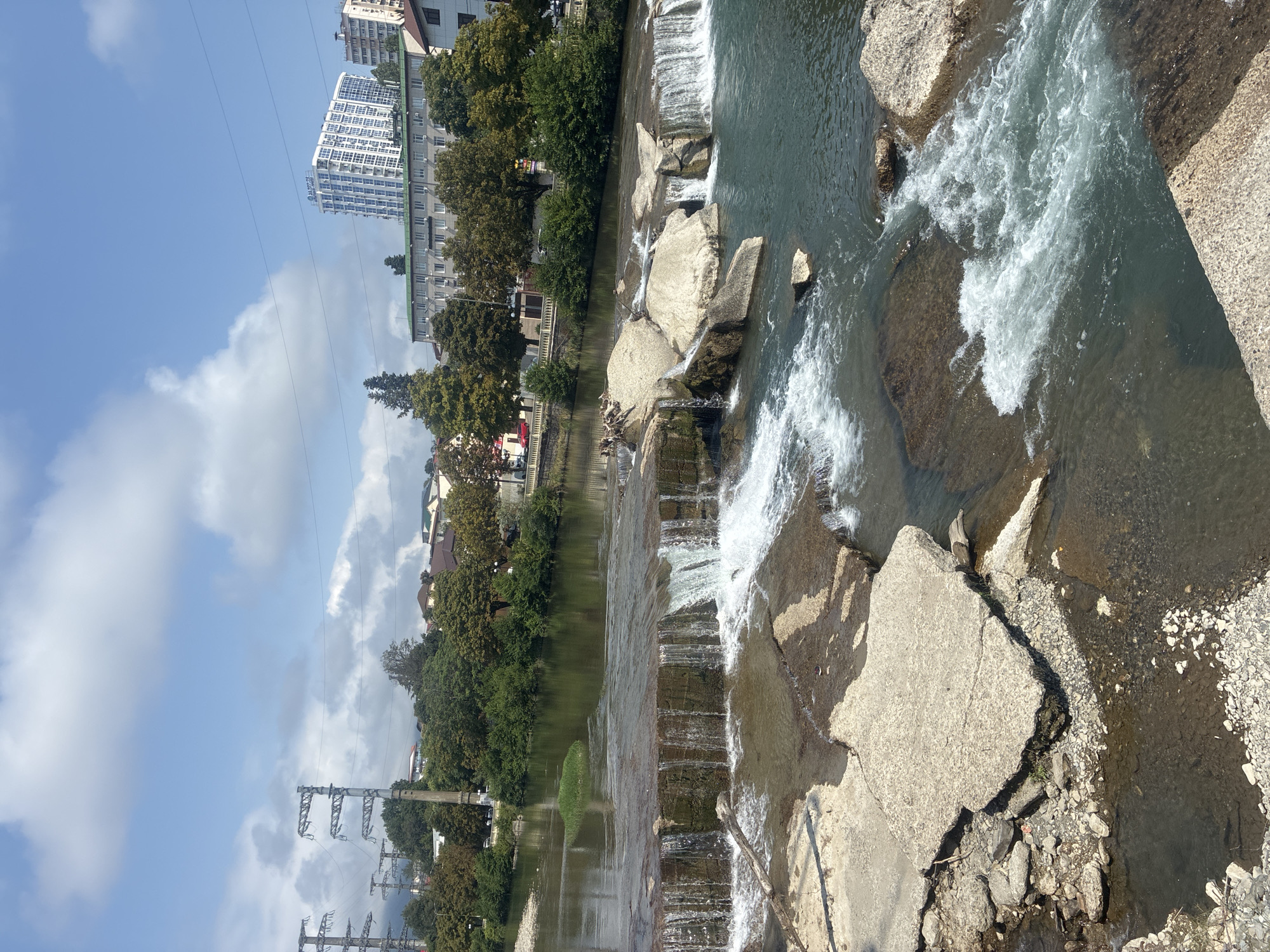 The image depicts a view of the Psou River in Sochi, Russia, showcasing a small waterfall. The foreground features the cascading water over concrete structures and natural rocks, with some debris visible on the banks. In the midground, the river flows calmly, with a small patch of green vegetation in the center. The background reveals a tree-lined embankment with various buildings, including a tall, modern apartment building. Power transmission towers are visible on the left side of the image, and the sky is partly cloudy. The scene is captured on a bright, sunny day.