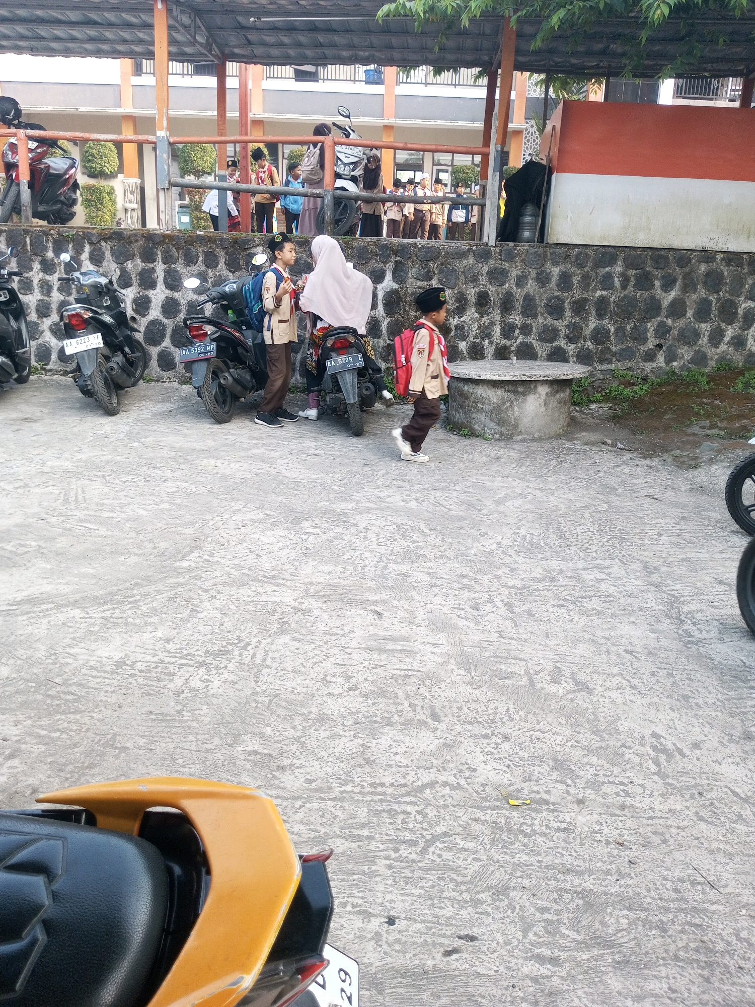 In Krasak, Indonesia, a group of students in school uniforms are gathered in an outdoor area near a building. Several motorcycles are parked on a concrete surface. In the foreground, a young student wearing a tan uniform, a black cap, and a red backpack walks to the right. Behind him, another student in a similar uniform and a pink hijab is interacting with someone out of frame. In the background, more students are visible, some standing in a line. The environment appears to be a school or educational facility with a stone wall and lush green foliage. The lighting suggests it is daytime.