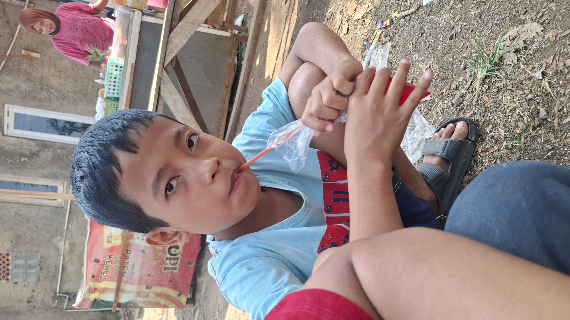A medium shot shows a young boy of Indonesian ethnicity, with dark hair and eyes, crouched on the ground, drinking from a plastic bag with a straw. He is wearing a light blue t-shirt with a red graphic on the front, dark shorts, and gray sandals. His legs are crossed in front of him, and his hands are holding the bag. The background includes a weathered building, a wooden cart with various items on it, and a woman in a pink shirt and headscarf standing behind the cart. The ground is covered with dirt and scattered debris. The scene appears to be outdoors, possibly in a marketplace or street in Cilaku, Indonesia.