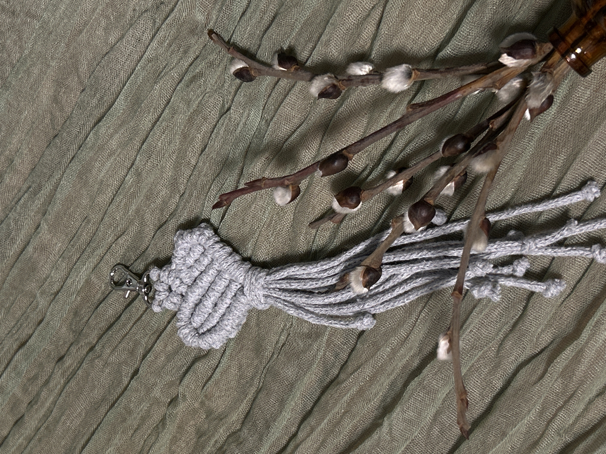 The image shows a gray, knotted macrame keychain with a silver clasp laying on a green, textured fabric. Next to it, there are several brown willow branches with white, furry buds. The top right of the image shows a portion of a brown vase. The scene appears to be indoors and lit by artificial light. There is no indication that the photo was taken in Saint Petersburg, Russia.