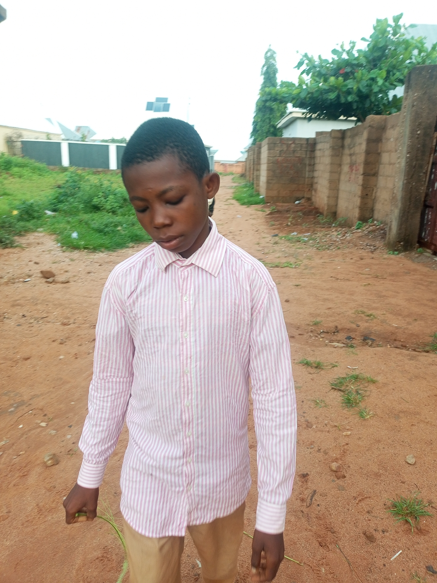 This media file is a medium shot of a young boy standing outdoors. He is wearing a light pink and white striped button-down shirt and tan pants. The boy is looking down, with his eyes closed or mostly closed, and appears to have a somber or thoughtful expression. He is holding a small bunch of green grass in his left hand.
The setting is a dusty, outdoor environment, likely in Tempure, Nigeria. The ground is reddish-brown dirt with scattered pebbles and patches of dry grass. In the background, there is a low, light-colored building with a dark fence and some greenery. Further back, a rough brick wall with concrete posts runs along the side of a path. There are also trees with lush green leaves and some red fruits visible behind the wall. The sky is overcast and bright, suggesting it is daytime, possibly in the morning or afternoon. There is no visible text in the image.