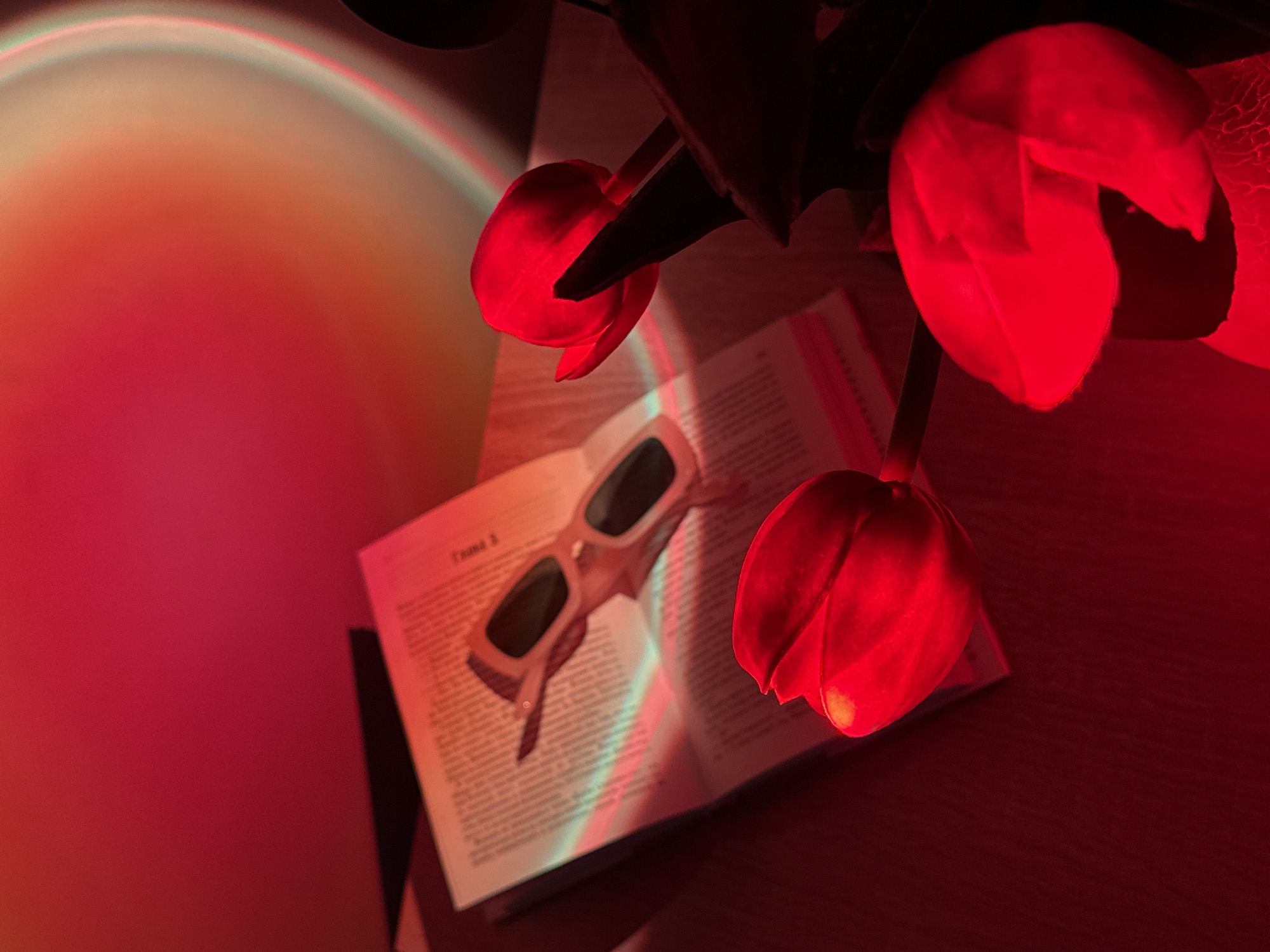 The image depicts a still life arrangement with red tulips, an open book, and sunglasses. The scene is bathed in a warm, reddish light that casts colorful reflections and shadows. A rainbow-like refraction of light, showing bands of green and blue, cuts across the book and sunglasses. The tulips are vibrant red and appear to be fresh, with their petals slightly unfurled. They are positioned to the right of the frame, with one in the foreground and two others further back.
An open book lies on a wooden surface, its pages filled with text. Placed on top of the book are a pair of white sunglasses with dark lenses. The sunglasses are angled, suggesting they were recently set down. The overall impression is one of quiet contemplation or a moment of leisure.
The lighting suggests it might be evening or a dimly lit indoor space, possibly enhanced by artificial light to create the dramatic color effects. The background is a blend of deep reds and oranges, with a soft, diffused glow in the upper left corner. The scene evokes a sense of warmth, creativity, and perhaps a touch of mystery due to the unusual lighting.
Visible text on the book appears to be in Russian, with "Глава 3" (Chapter 3) clearly legible. The setting is intimate and personal, creating a mood that is both visually striking and emotionally resonant.