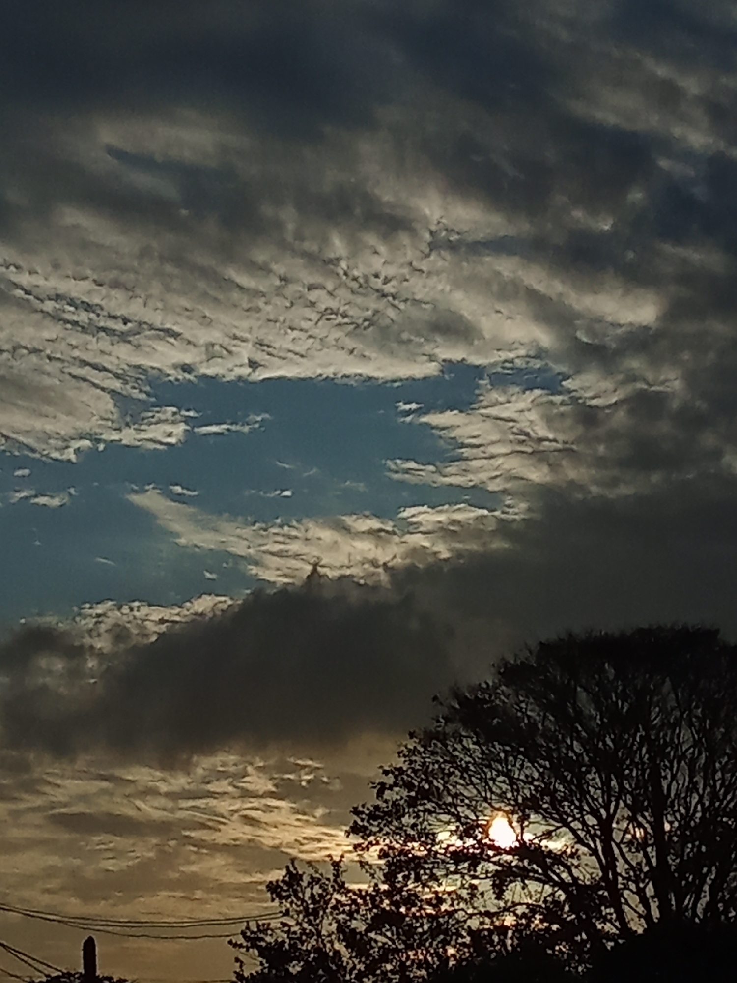 The image depicts a dramatic sky above what appears to be the city of Juan Griego, Venezuela, characterized by a mix of textured clouds. In the upper portion, thin, striated clouds are visible against patches of blue sky. The lower portion of the sky is filled with darker, heavier cloud formations. The silhouetted outline of a tree occupies the bottom right of the image, partially obscuring the setting or rising sun which is visible as a bright spot through its branches. A distant silhouette of what appears to be a utility pole is visible to the bottom left.